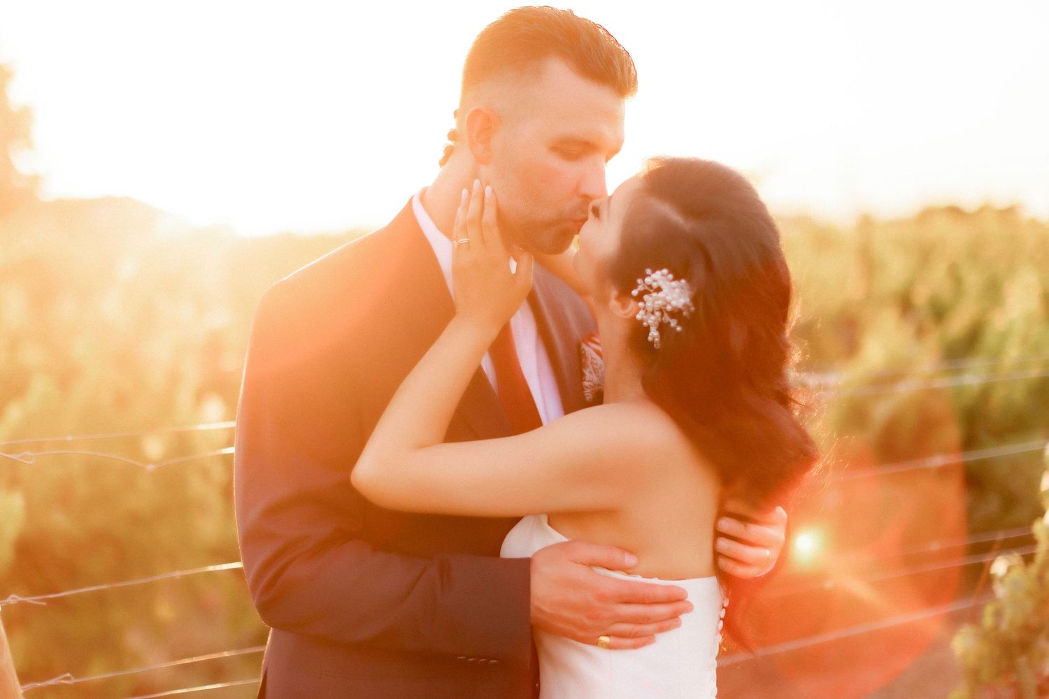 Couple embraces and kisses at sunset in golden field with glowing sun rays illuminating their silhouettes.