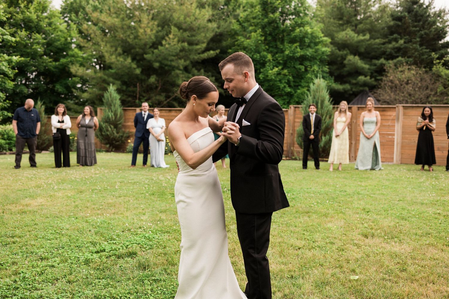 A couple shares their first dance at an outdoor wedding ceremony while guests watch from a distance on a green lawn.