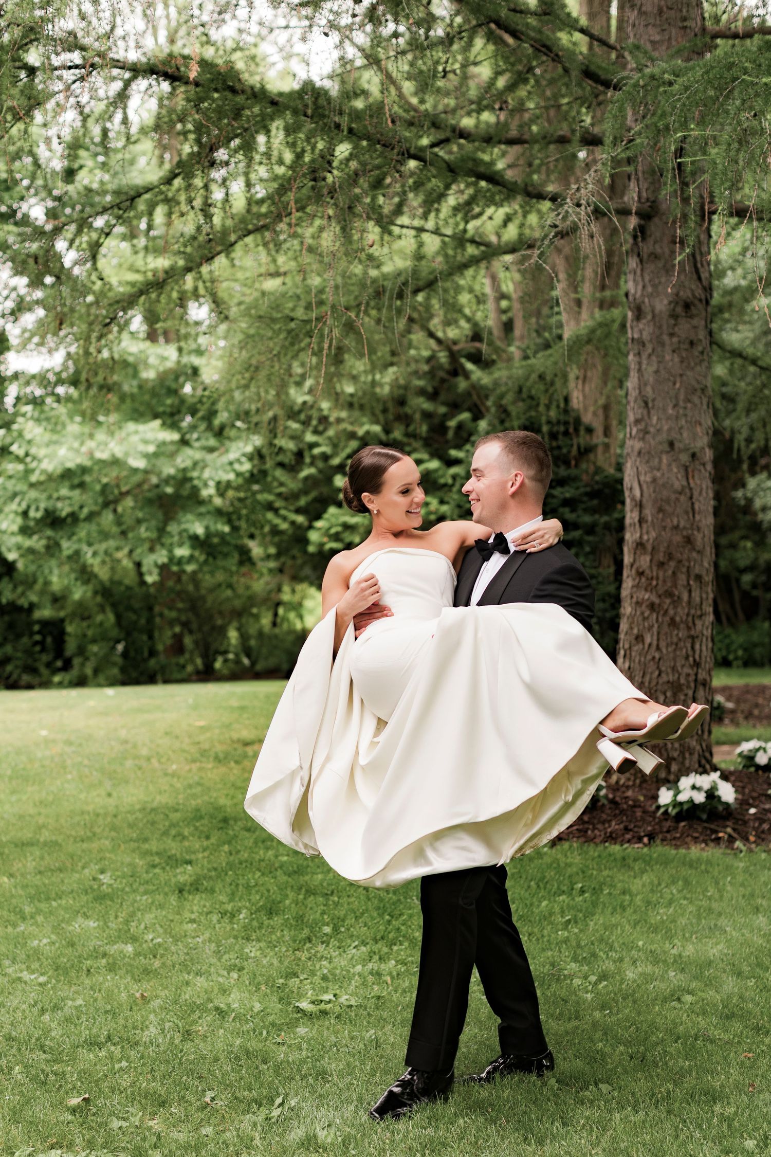 Groom carries bride in traditional pose surrounded by towering evergreen trees.