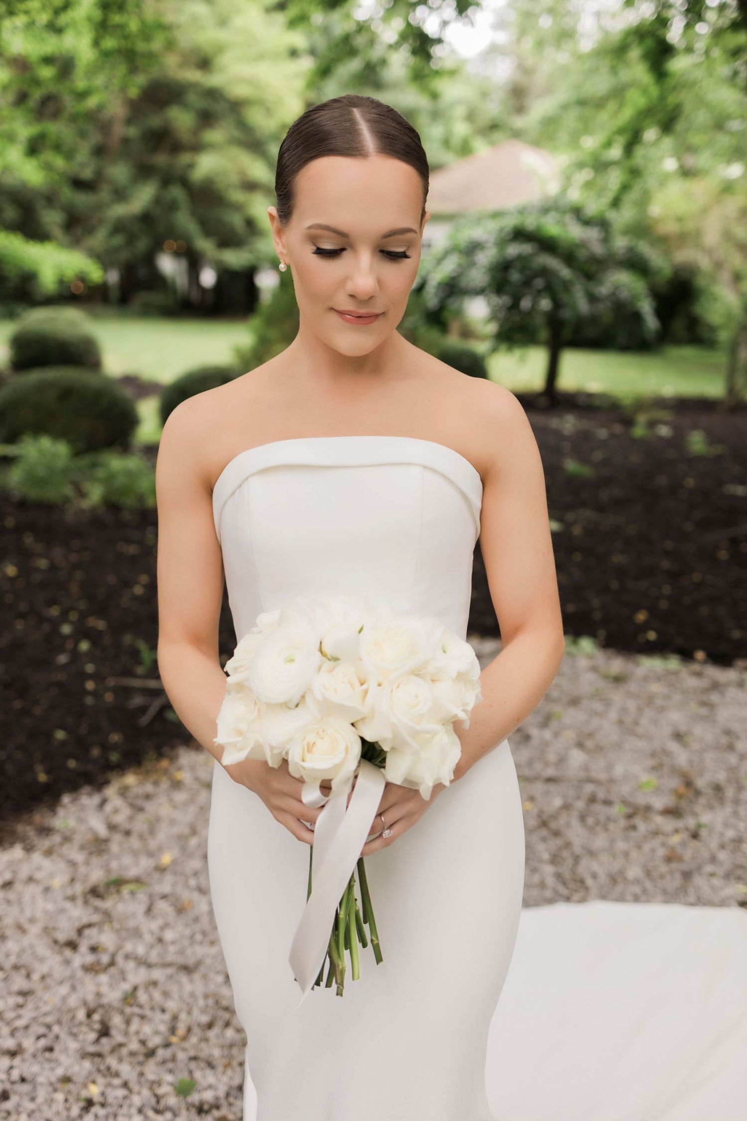 A minimal white wedding gown displayed in an outdoor garden setting with lush greenery.