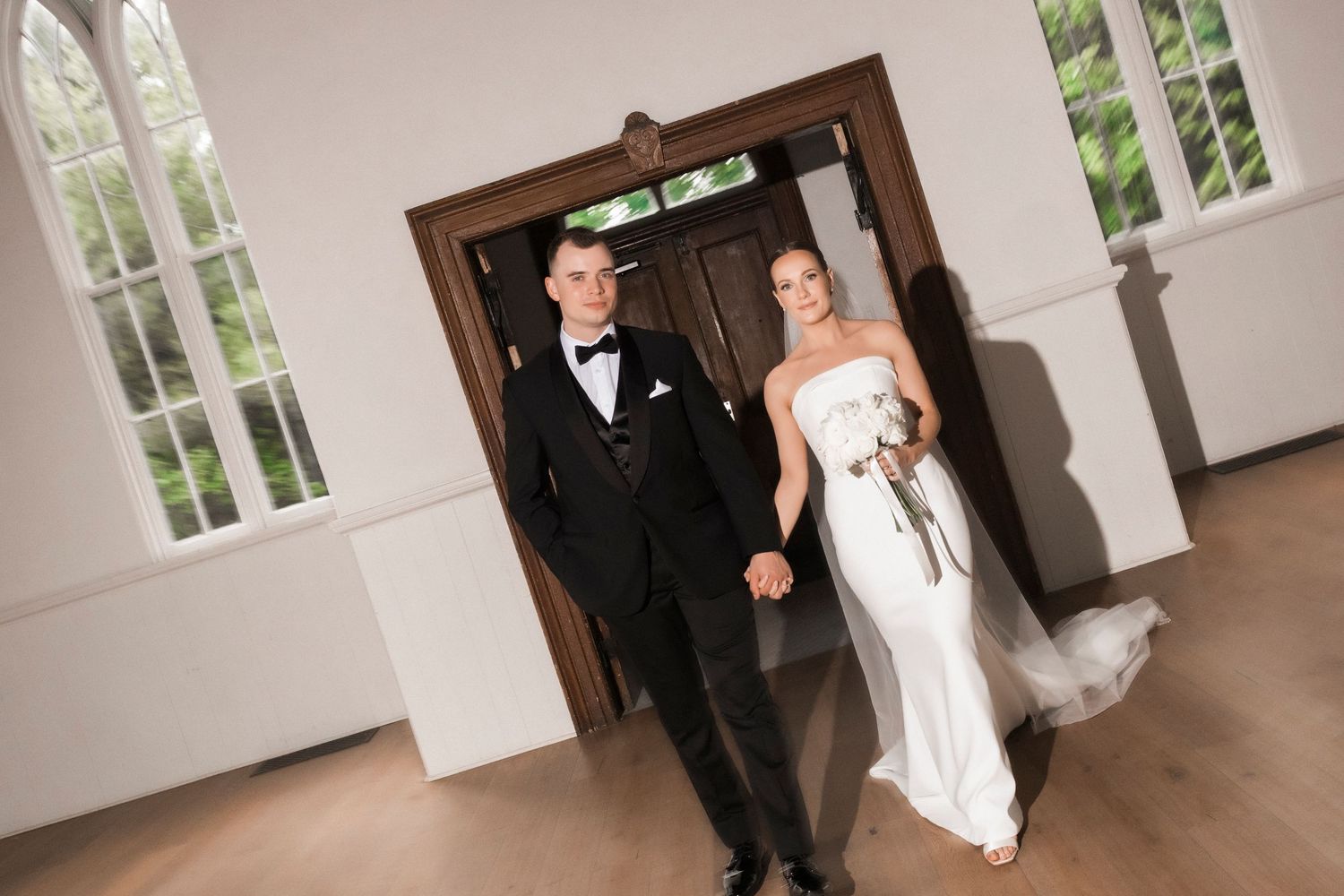 A wedding couple dances gracefully in a bright white room with arched windows and hardwood floors.