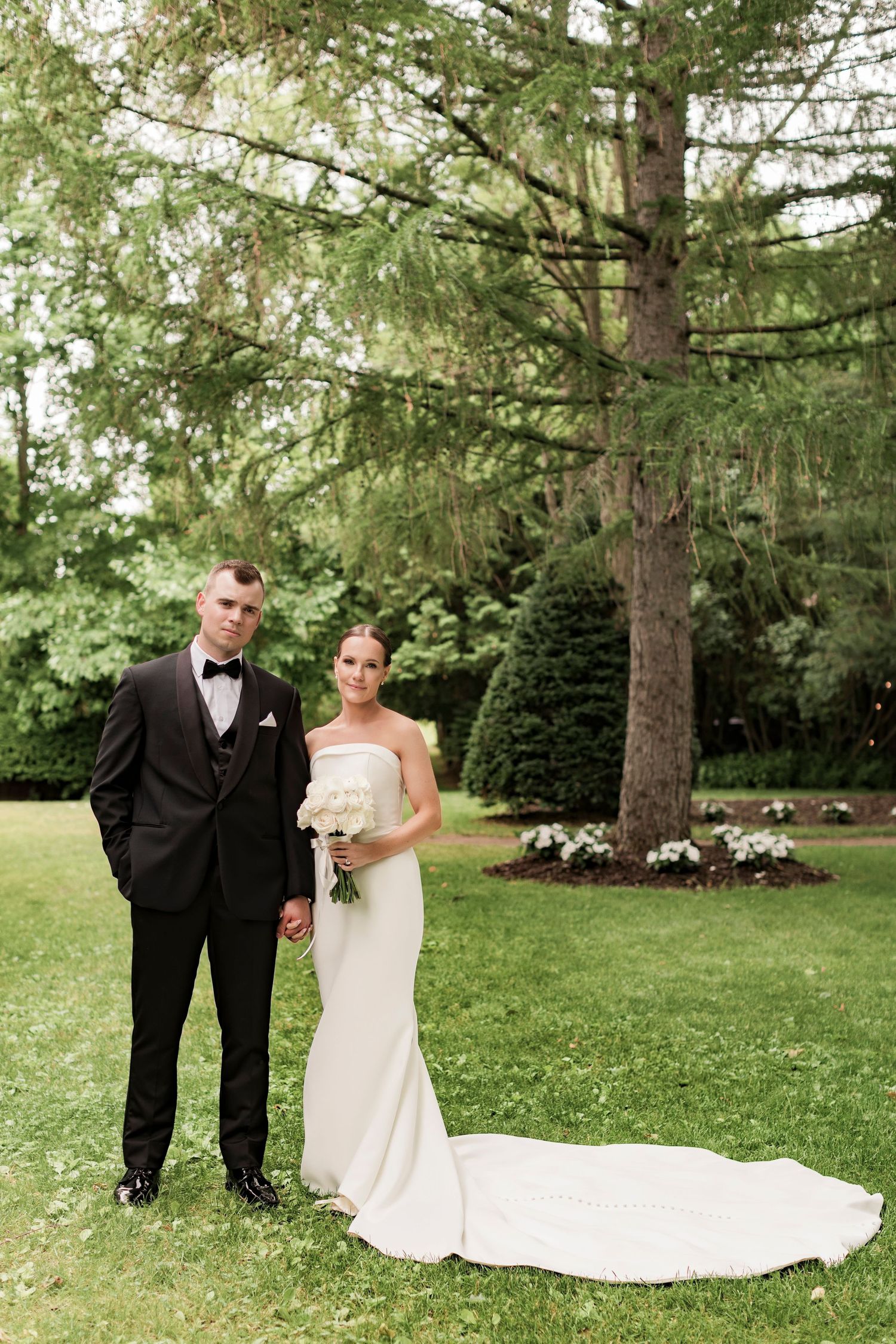 Formal wedding portrait of a couple standing beneath tall pine trees on a landscaped lawn.