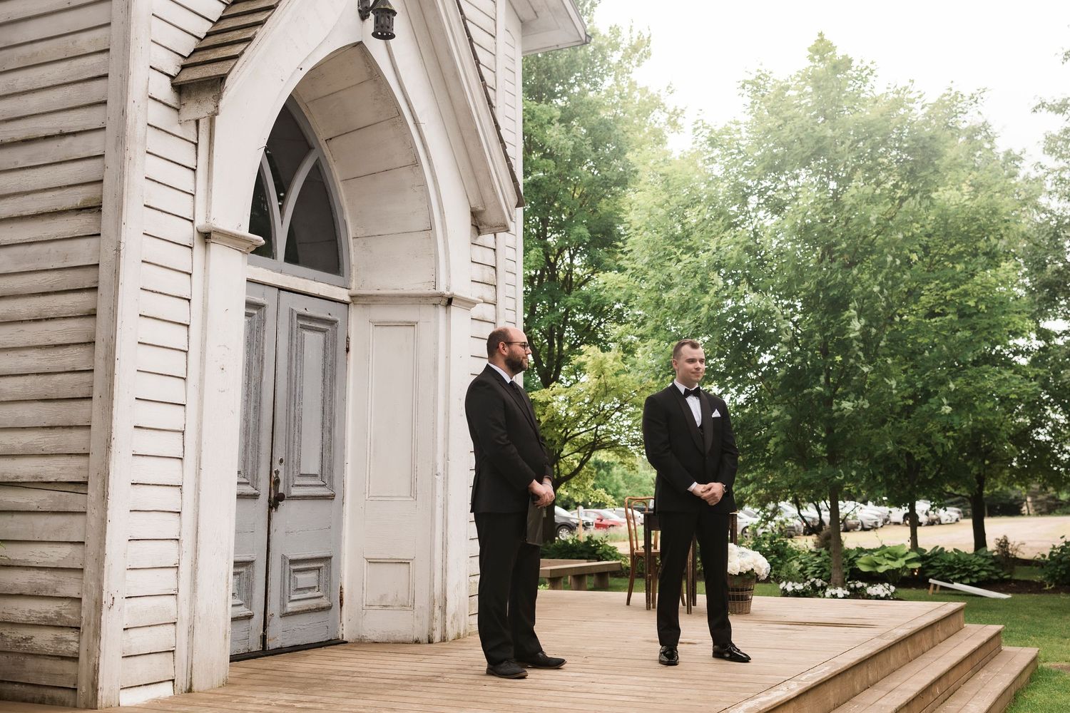 Two men in formal black suits stand at entrance of white church with arched doorway and leafy trees in background.