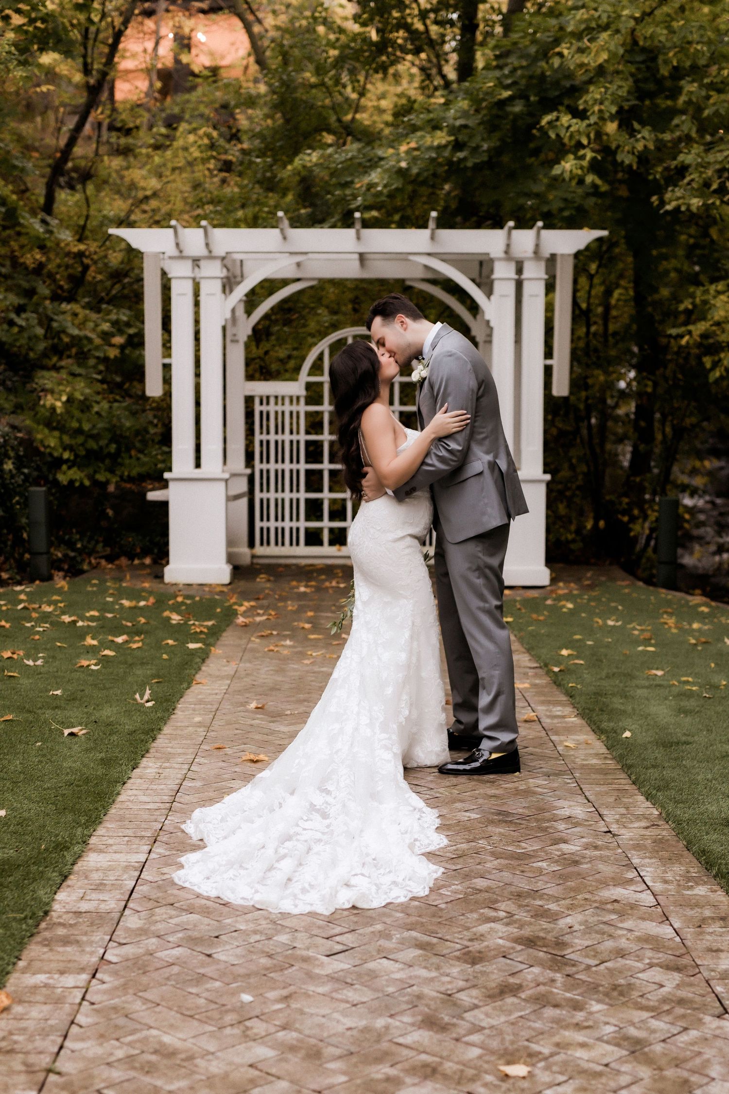 Romantic kiss under white garden arbor with bride's lace train extending down stone path.