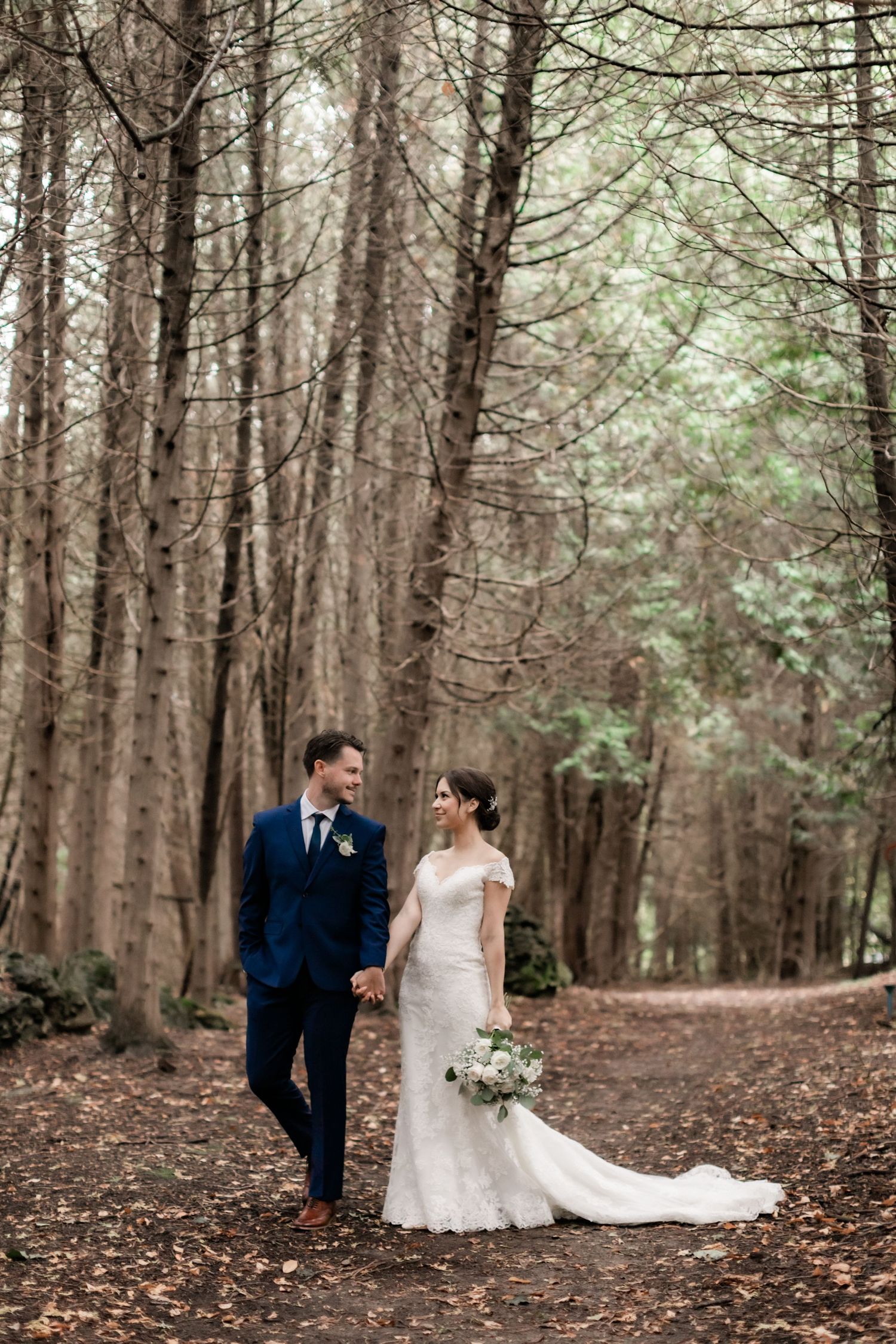 Bride in flowing lace gown and groom walking through a forest path lined with tall trees.