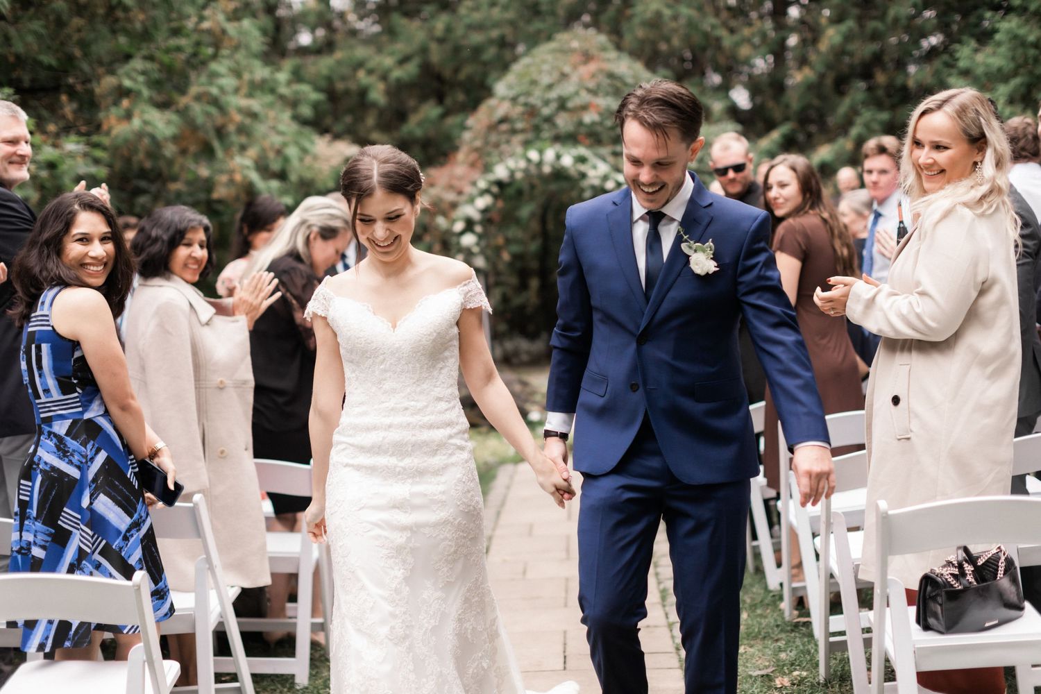 Newlyweds walk down the aisle at an outdoor garden ceremony surrounded by guests in elegant attire.