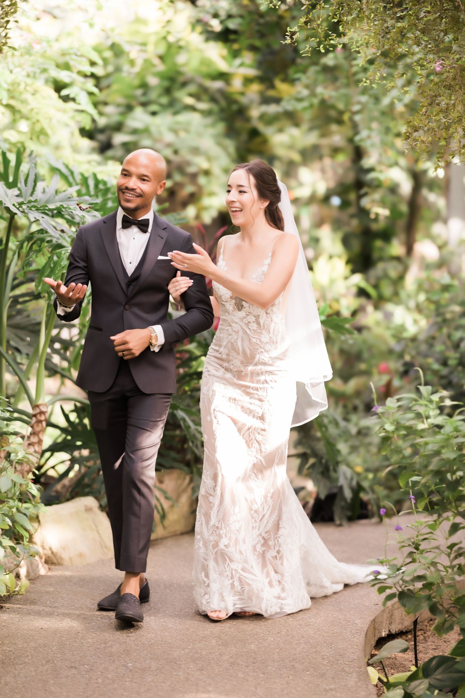 Wedding couple laughing while walking together on garden path surrounded by lush greenery.