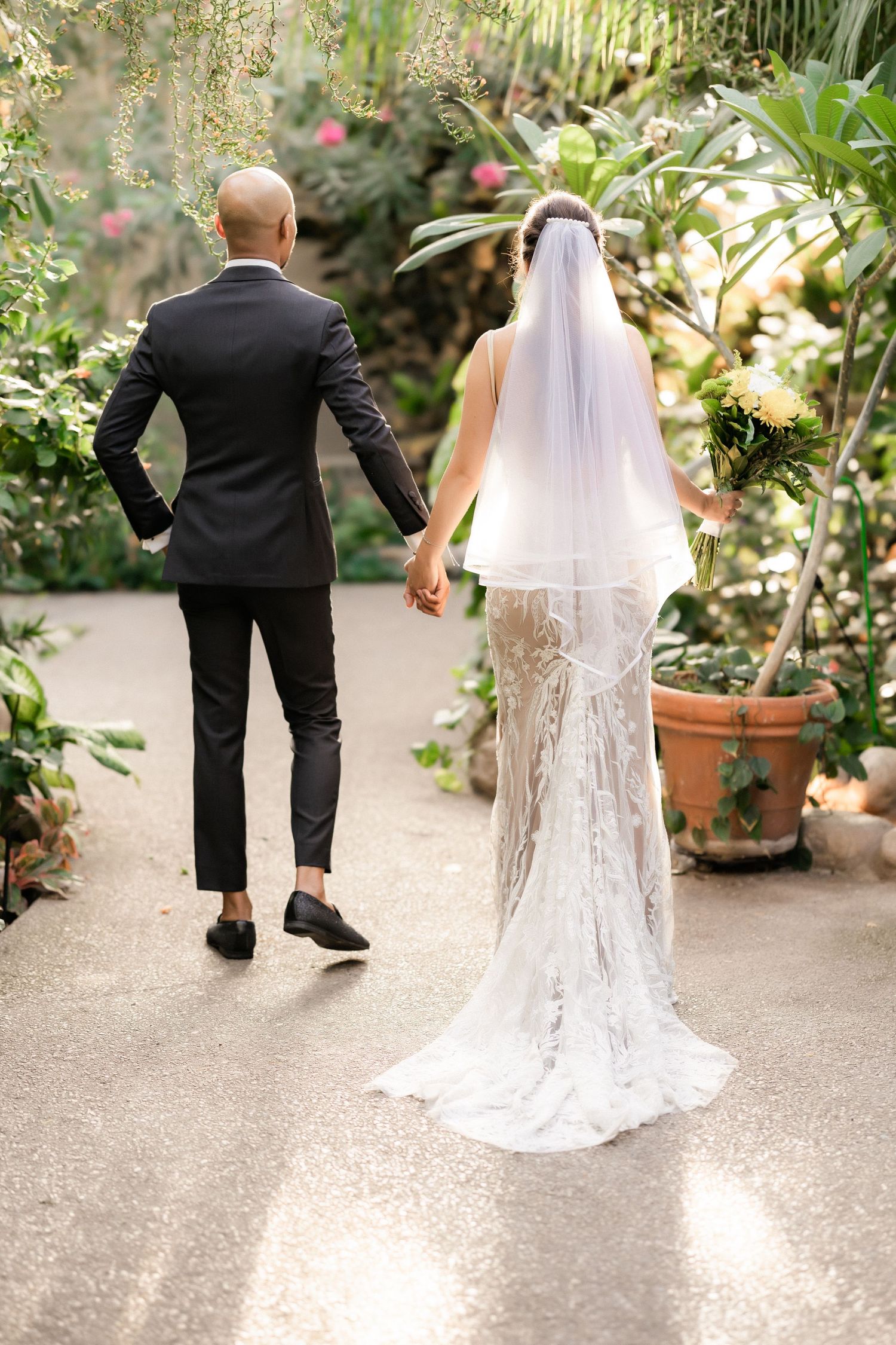 A wedding couple walks hand in hand down a garden path surrounded by tropical plants.