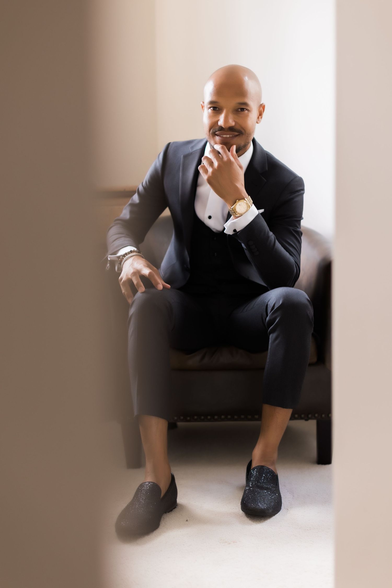 Dapper figure in formal black tuxedo poses contemplatively in dark leather armchair.
