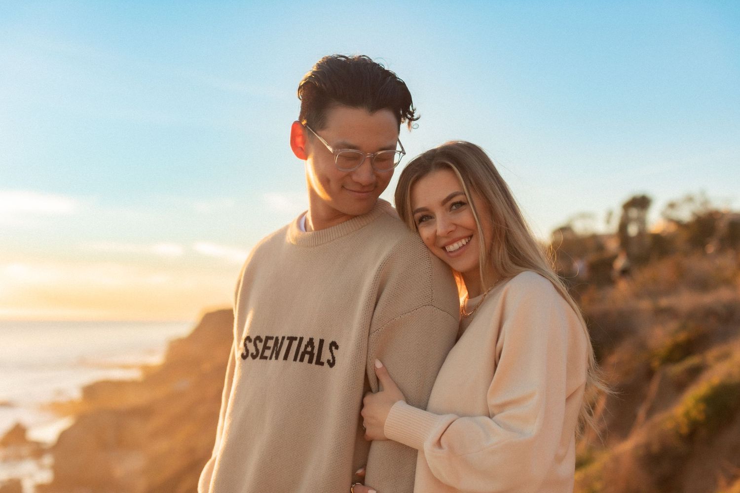 A couple embraces during sunset on a coastal cliff wearing casual sweaters against an ocean backdrop.