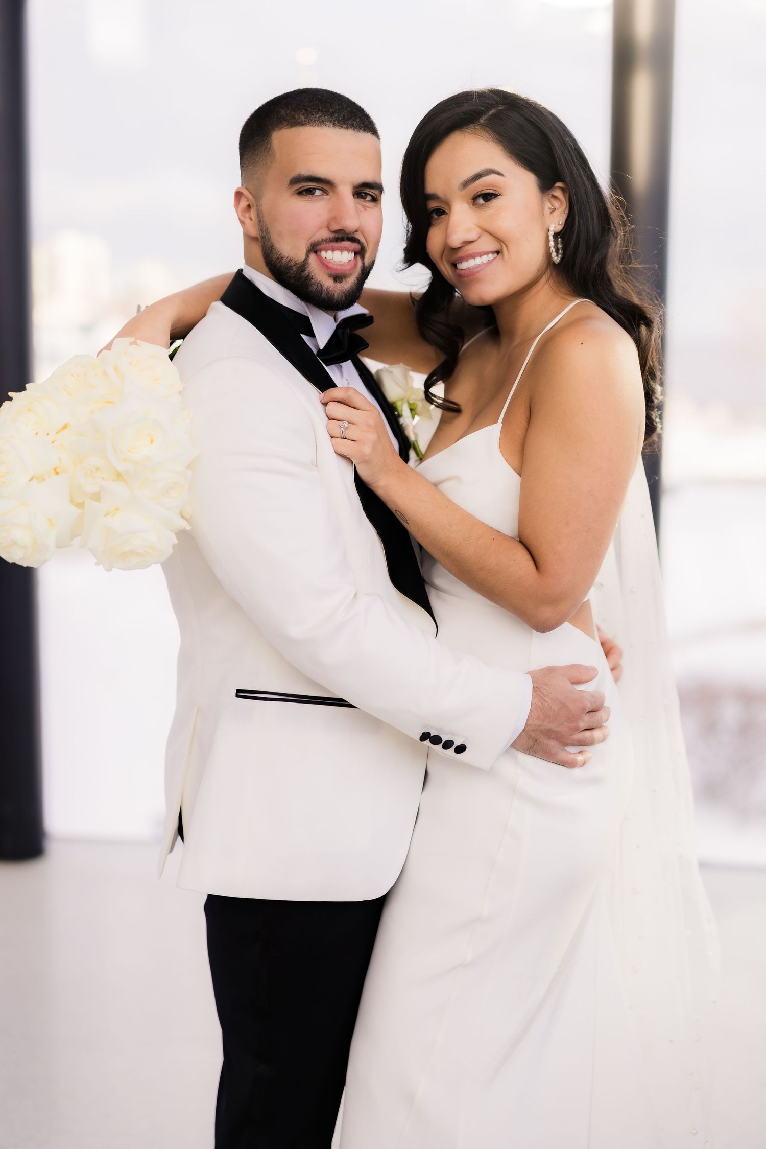 A joyful couple in matching white attire pose with a cream bouquet at their wedding.