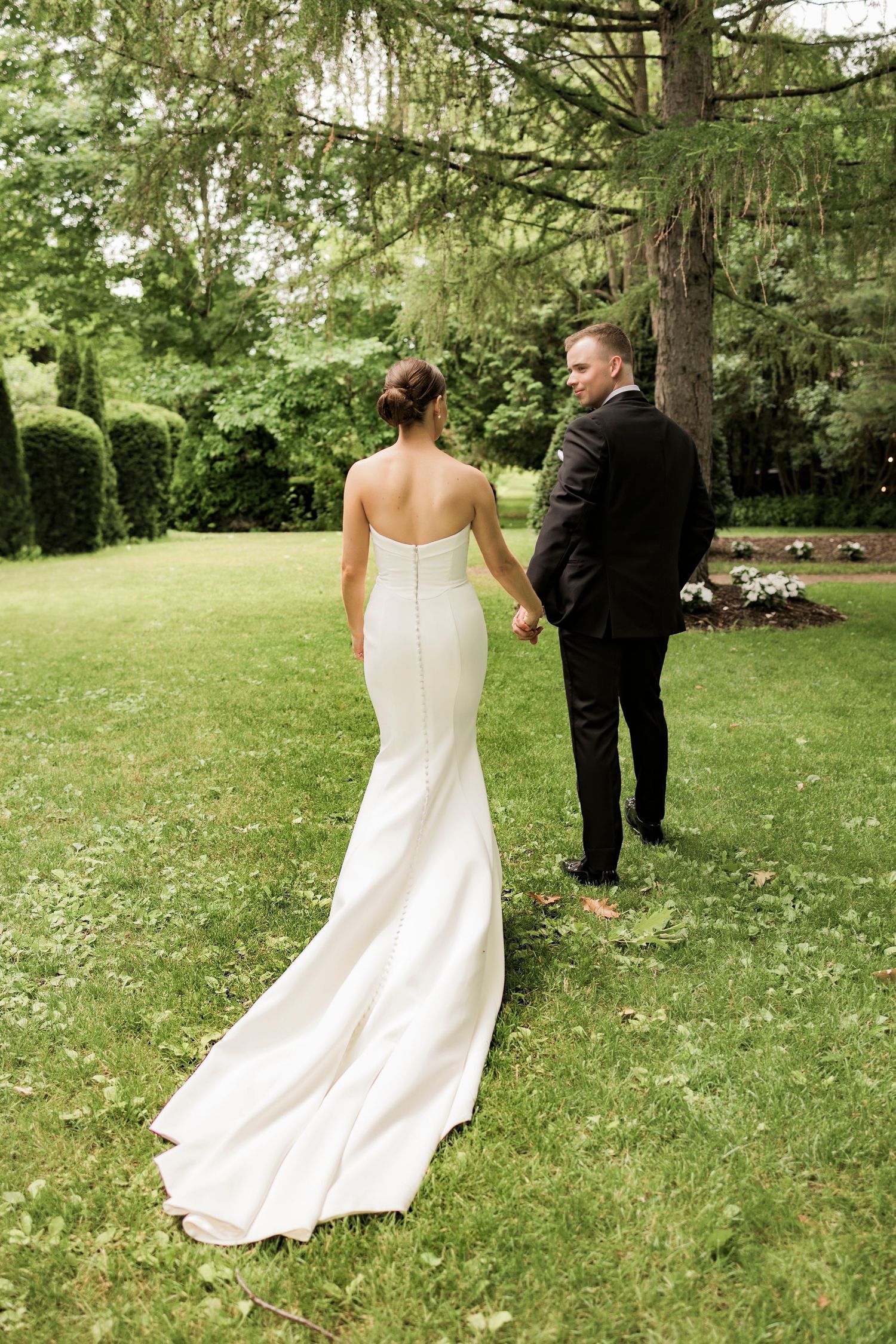 Elegant bridal couple holding hands while walking through lush garden landscape.