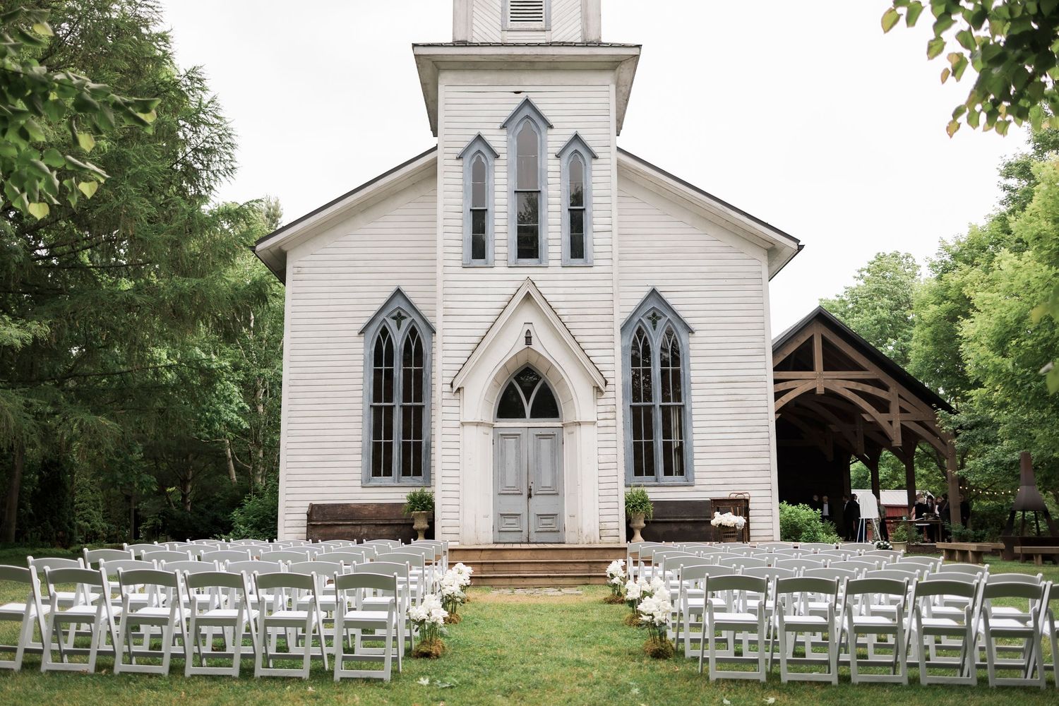 A rustic white church with Gothic windows sits amidst trees with white chairs set up for an outdoor wedding ceremony.