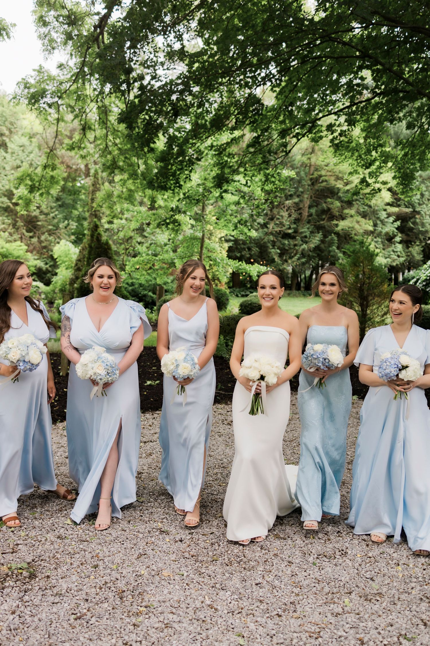 Wedding party in light blue bridesmaid dresses poses outdoors holding white bouquets on gravel path with greenery backdrop.