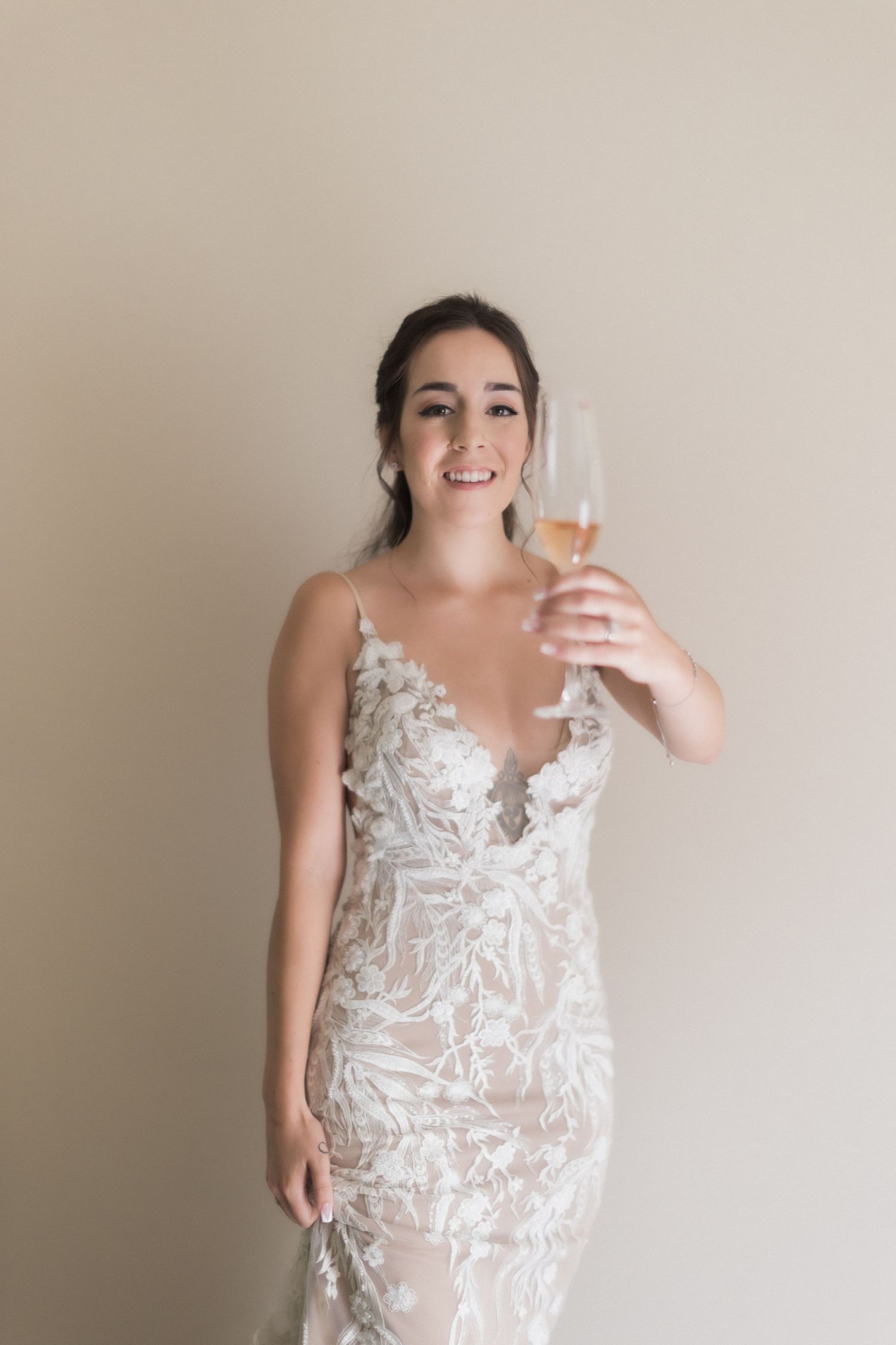 Someone in an elegant white lace wedding dress holds up a champagne glass against a neutral wall.