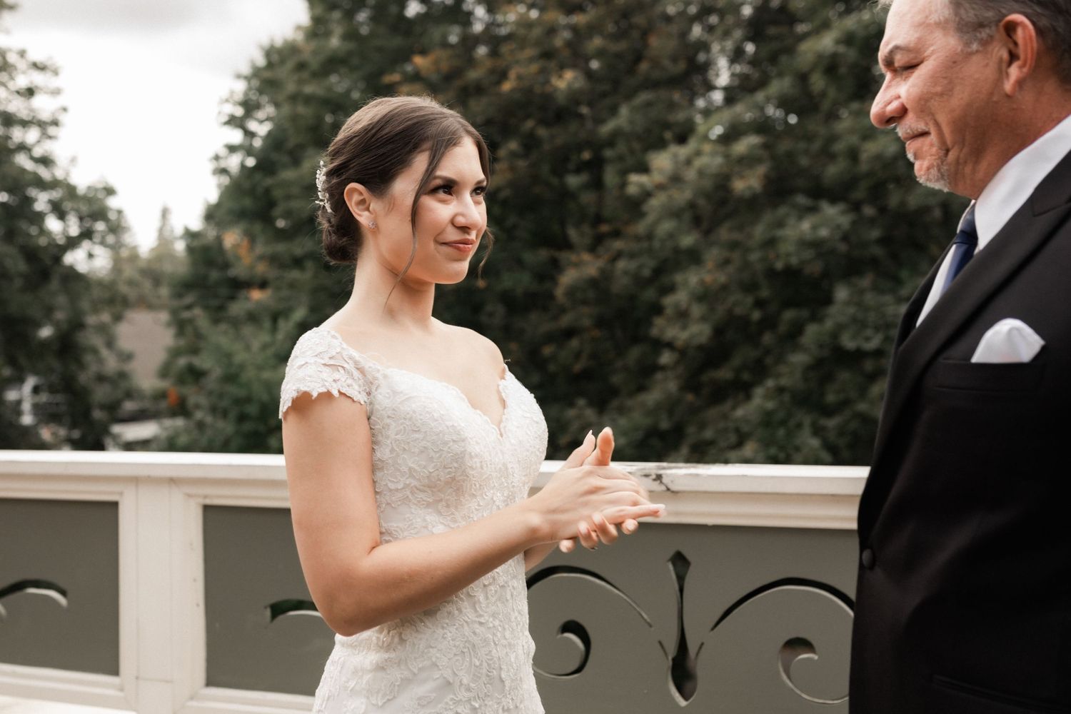Bride in lace cap sleeve wedding dress shares emotional moment with man in black suit on outdoor terrace.