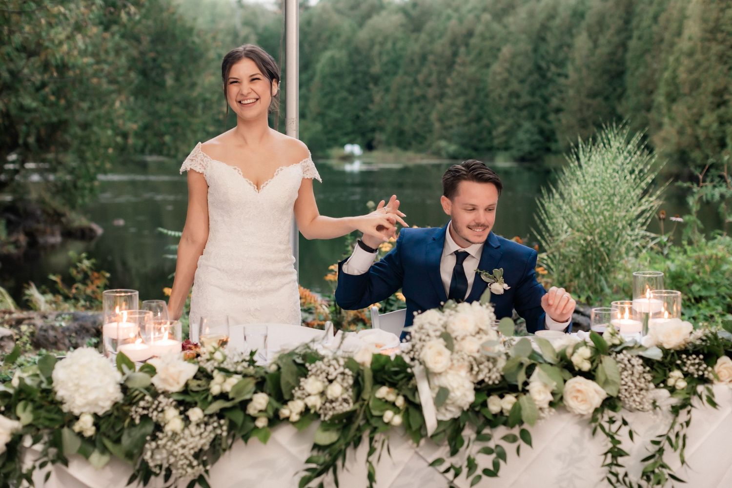 A wedding couple stands behind their reception table decorated with lush white flowers.