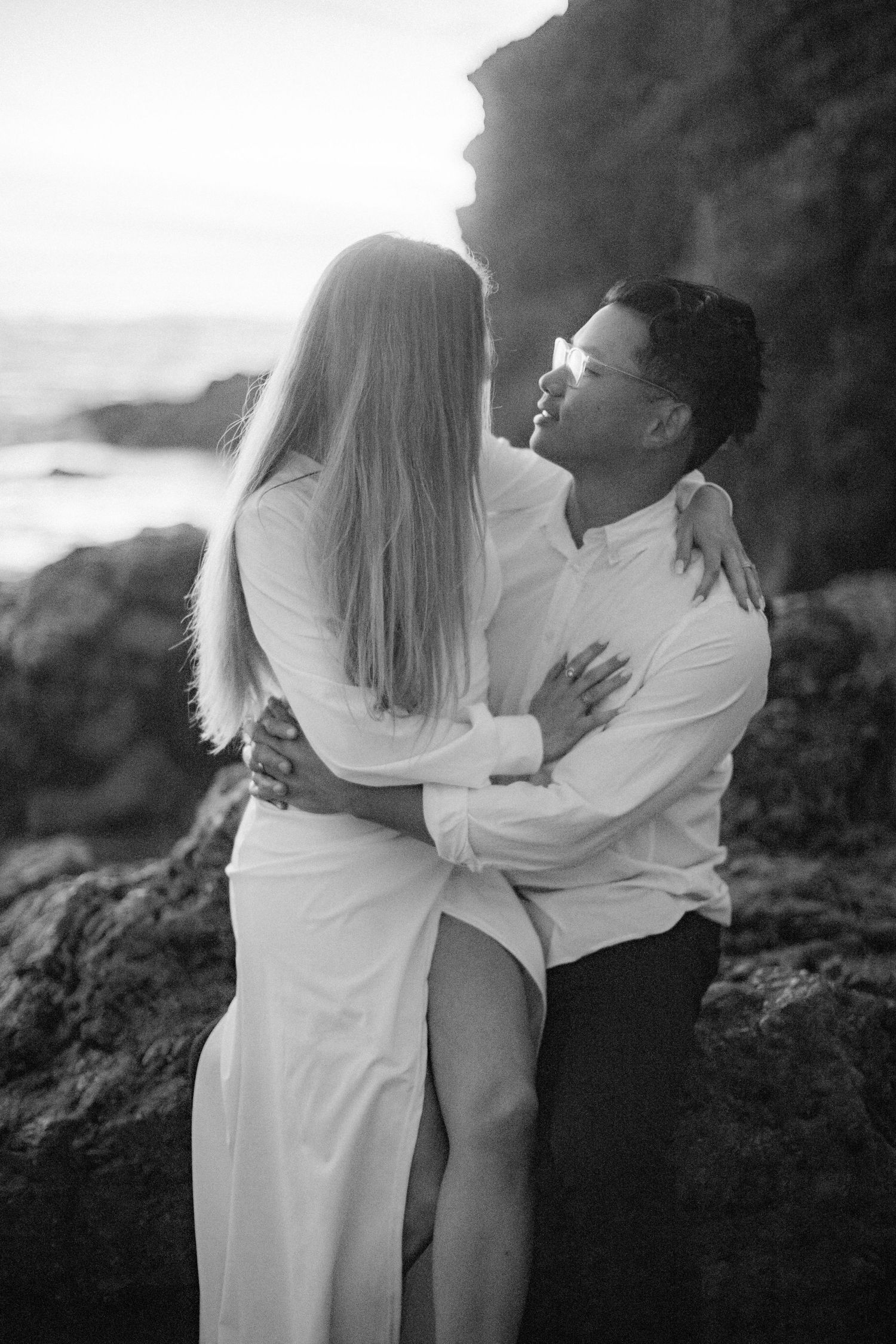 A romantic couple in white outfits embracing on coastal rocks during a black and white photoshoot.