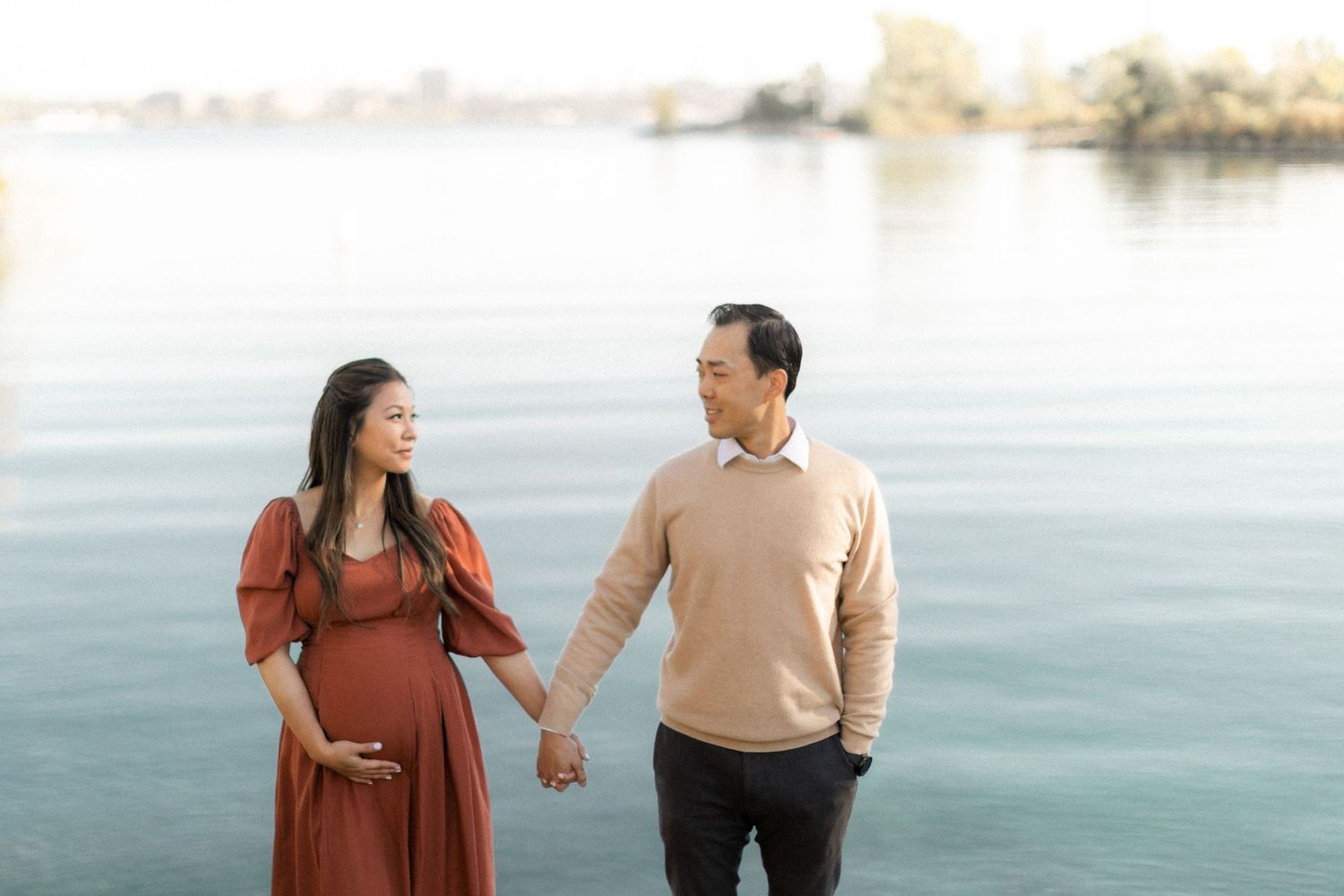 Expectant couple hold hands by serene lake, wearing beige sweater and rust dress.