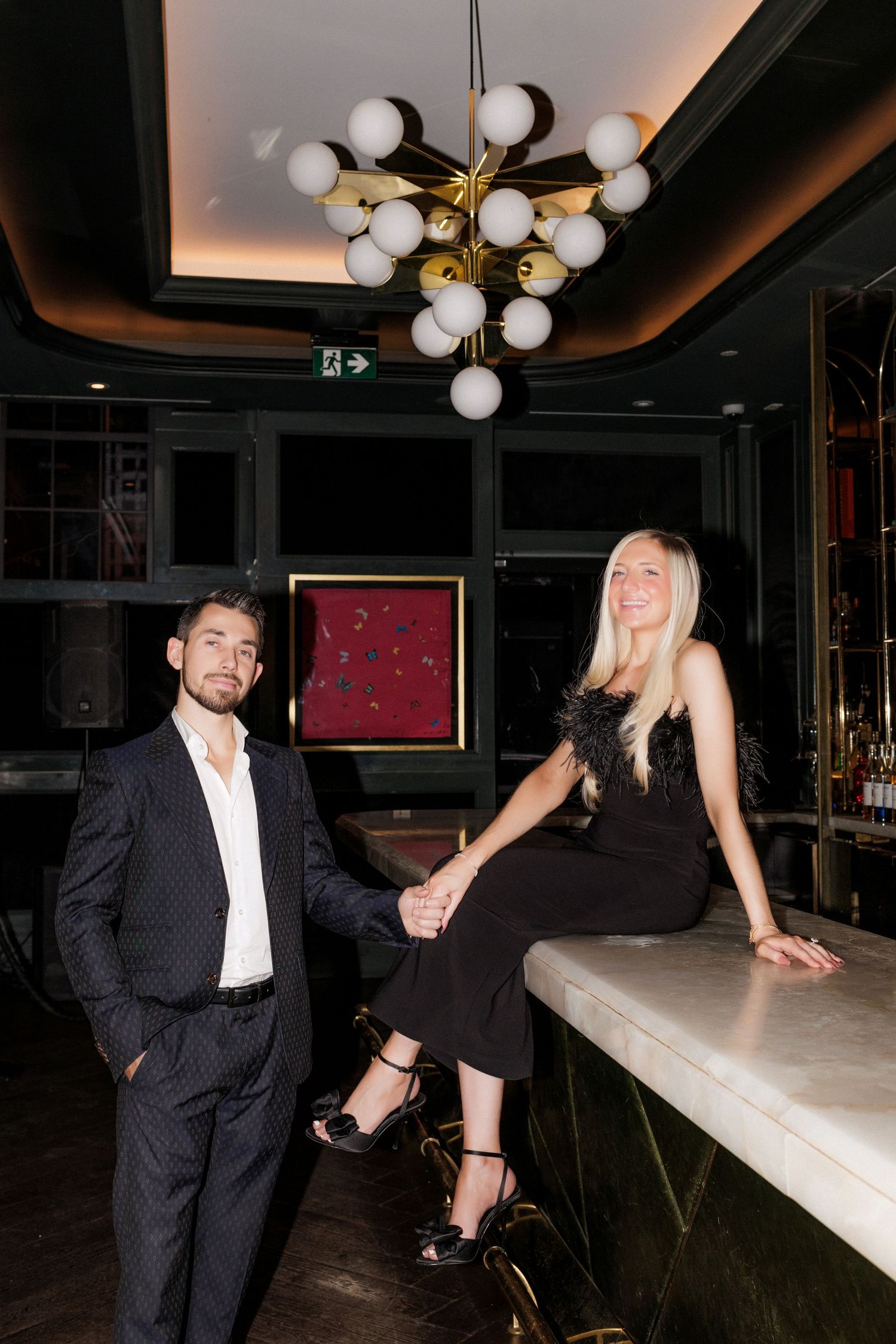 Elegant couple in formal attire posing at an upscale bar with spherical light fixture overhead.
