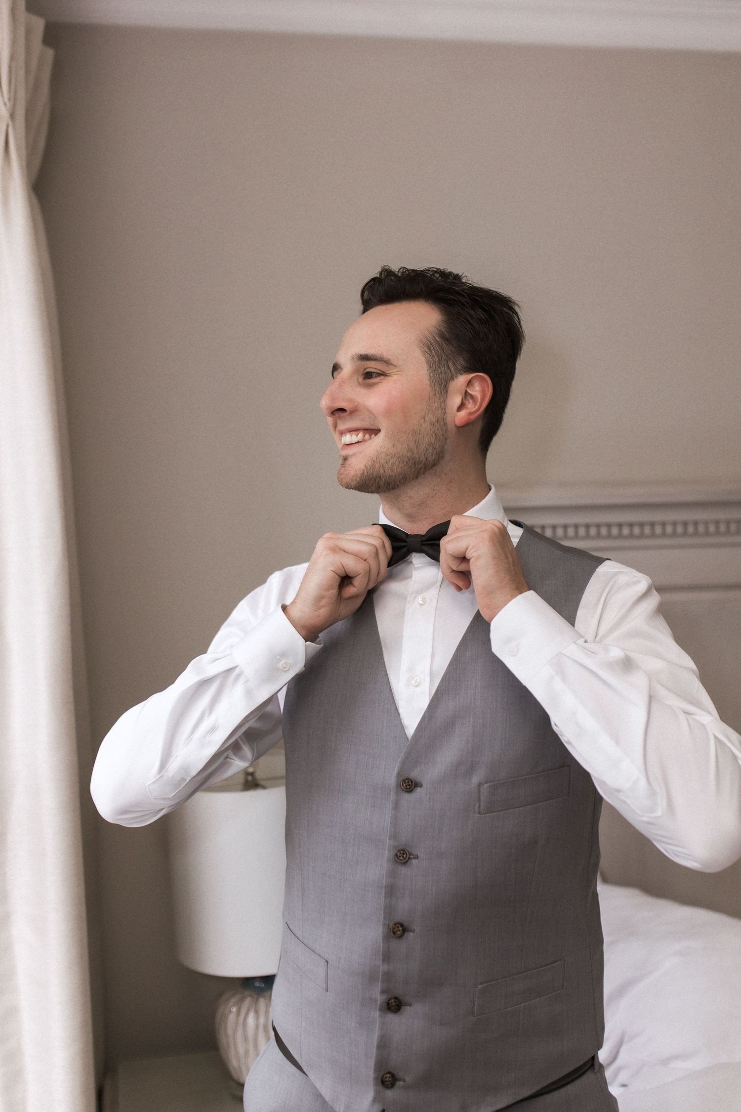 Groom smiles while adjusting his bowtie in elegant grey vest and white shirt.