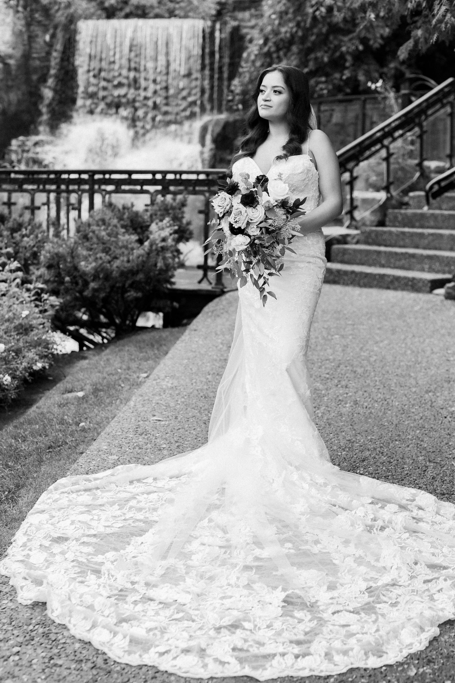Bride in lace gown with flowing train poses elegantly by waterfall in black and white portrait.