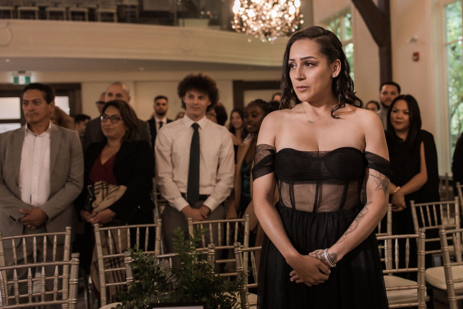 Wedding guest in black formal dress standing solemnly during indoor ceremony.
