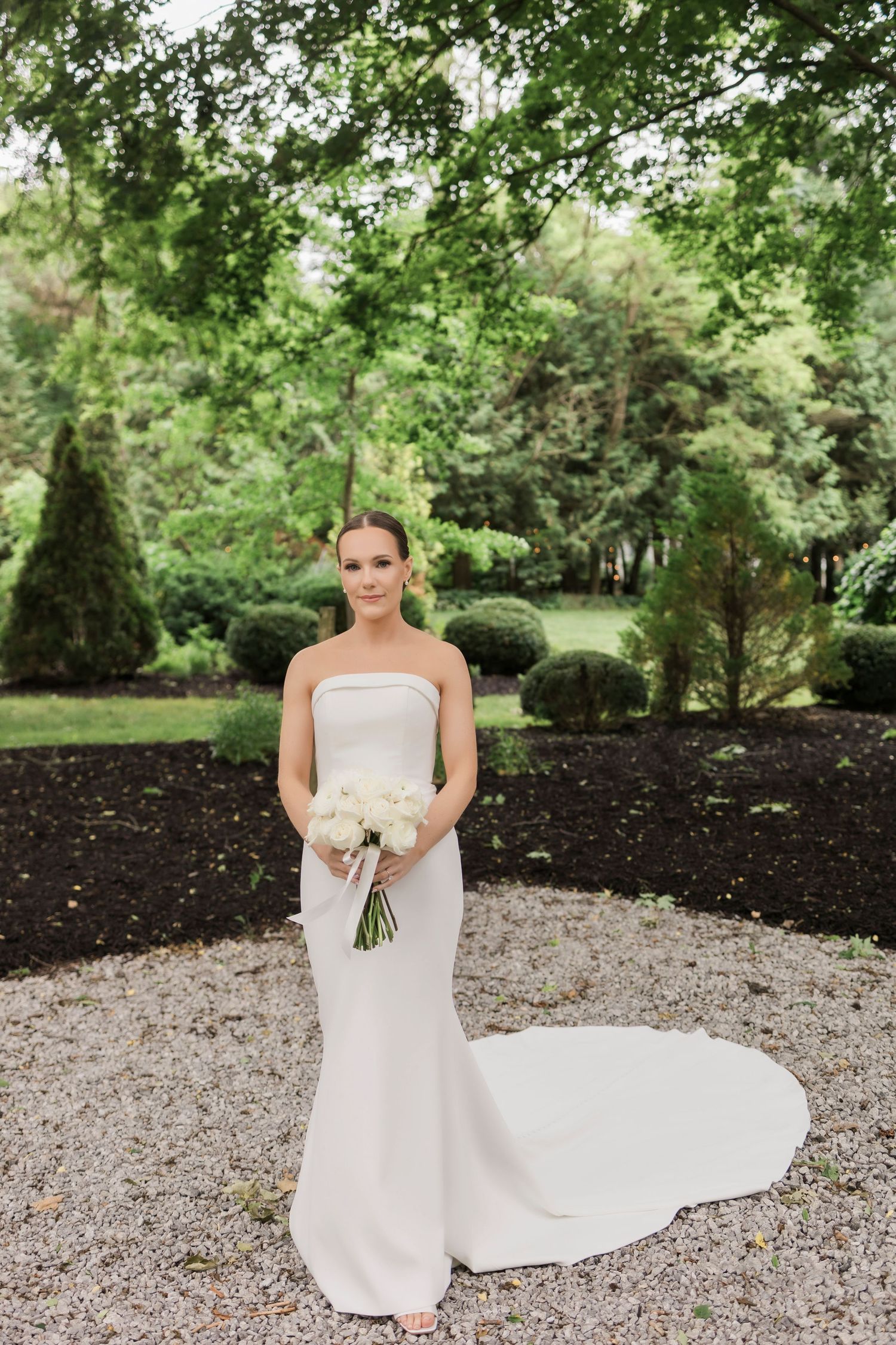 Elegant strapless wedding gown with fitted silhouette and white bouquet in a manicured garden setting.