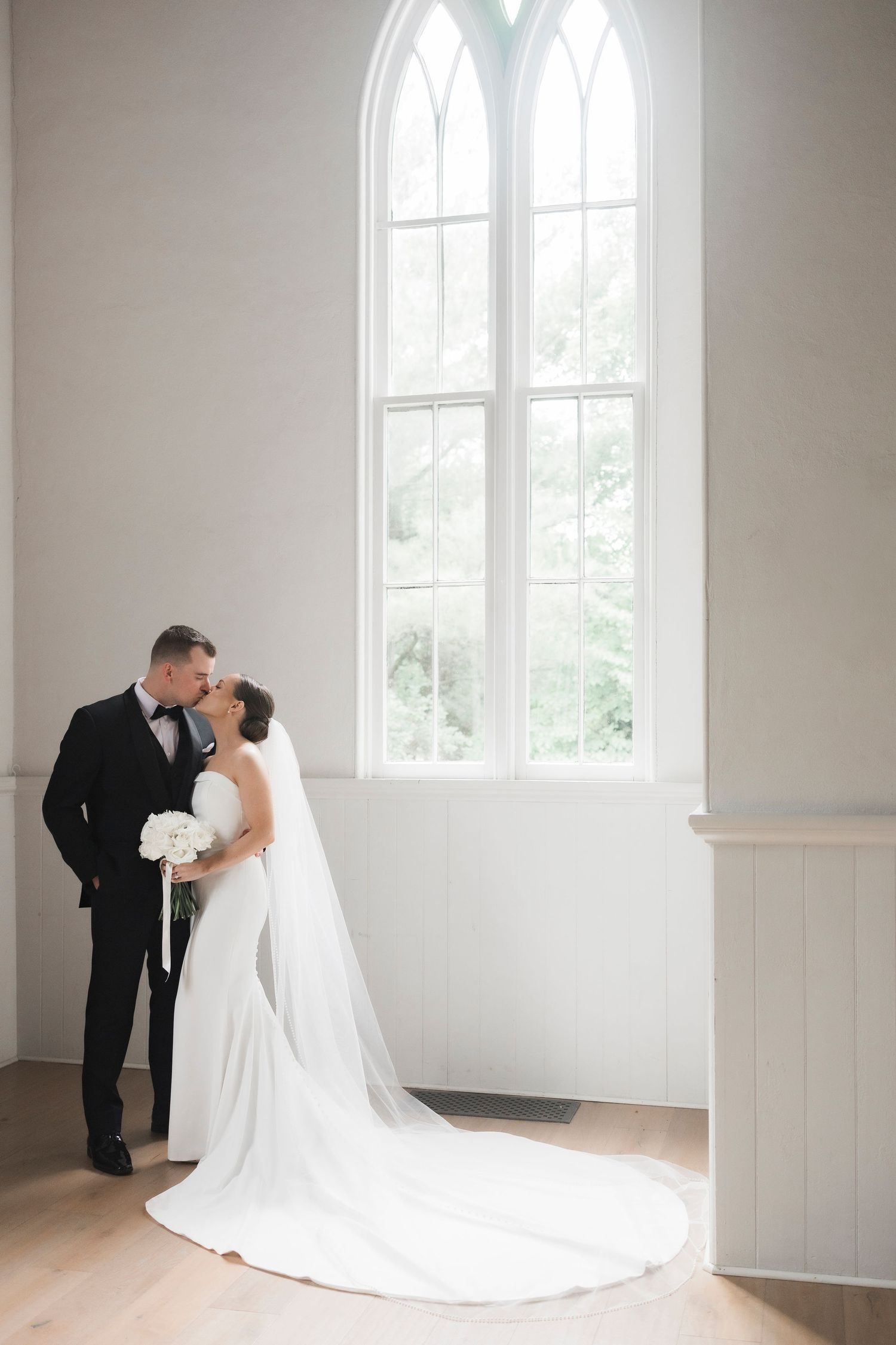 A romantic wedding moment with couple standing by ornate church window in elegant attire and long white train.
