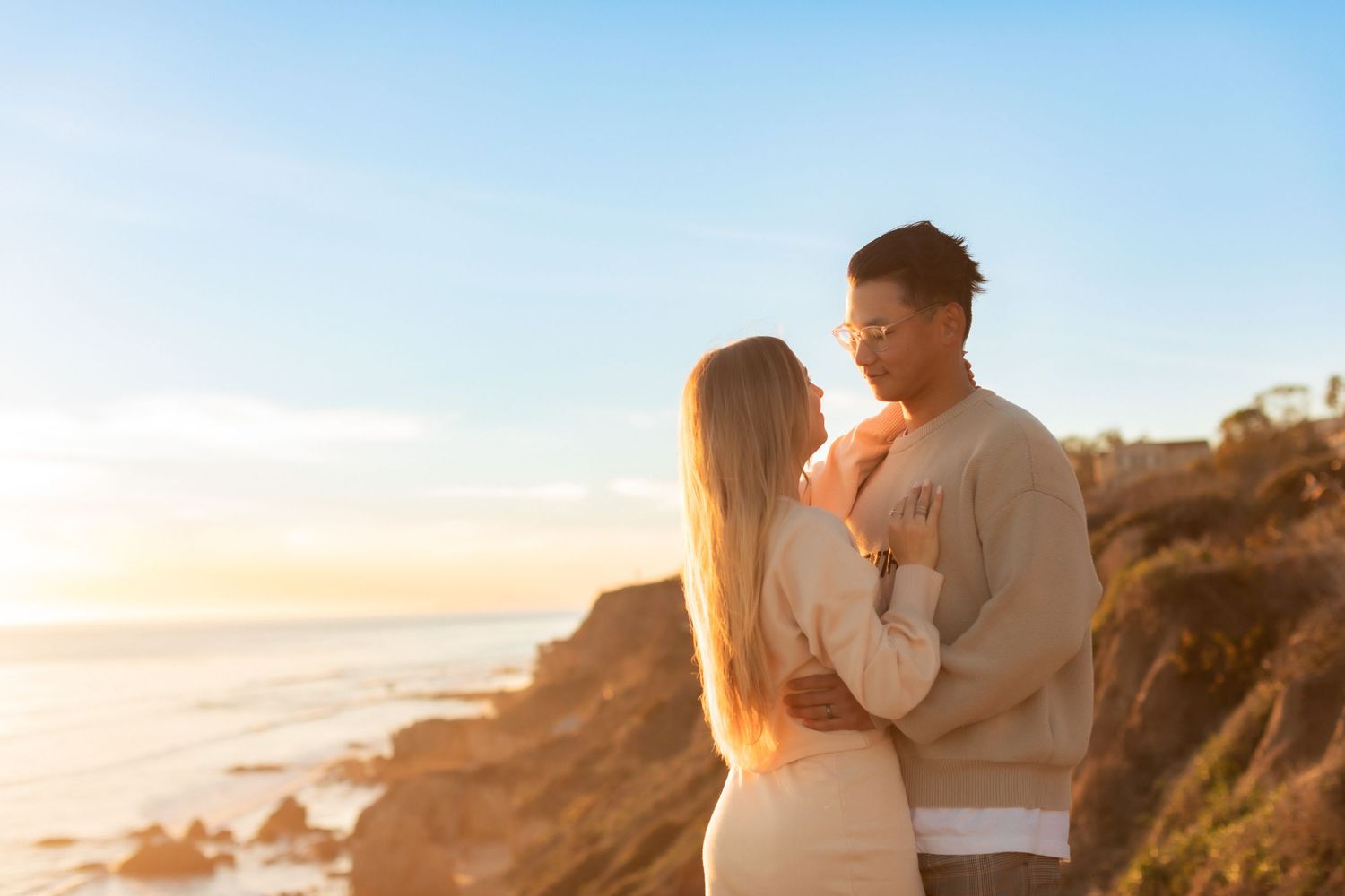 Couple in matching neutral outfits embracing against sunset ocean view.
