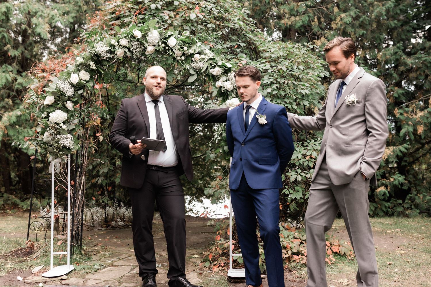 Groomsmen in suits share a lighthearted moment under flowering trees at an outdoor ceremony.