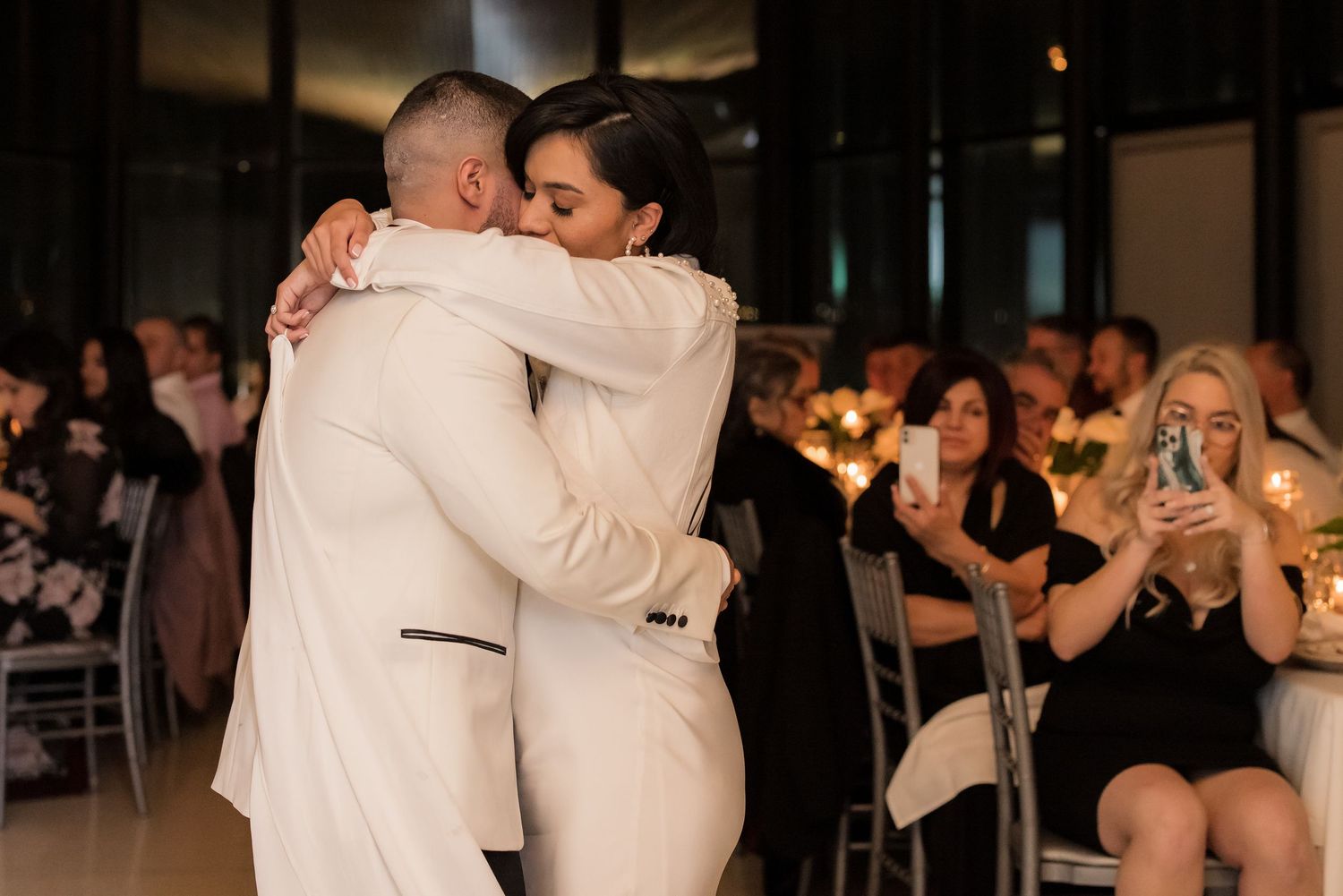 A couple in elegant white attire share a dance while wedding guests watch and take photos.