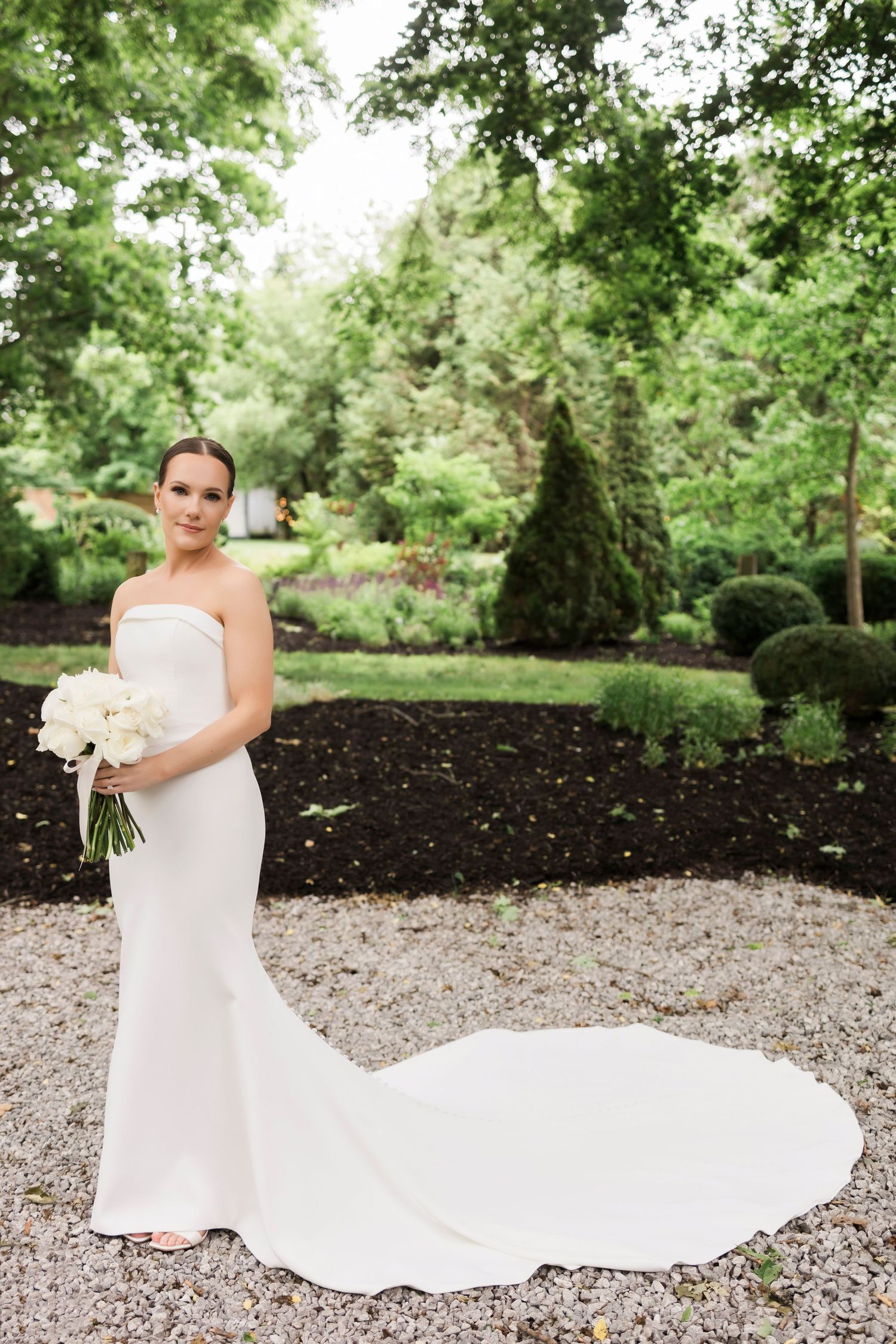 Elegant bride in strapless white gown with long train poses in garden setting holding white bouquet.