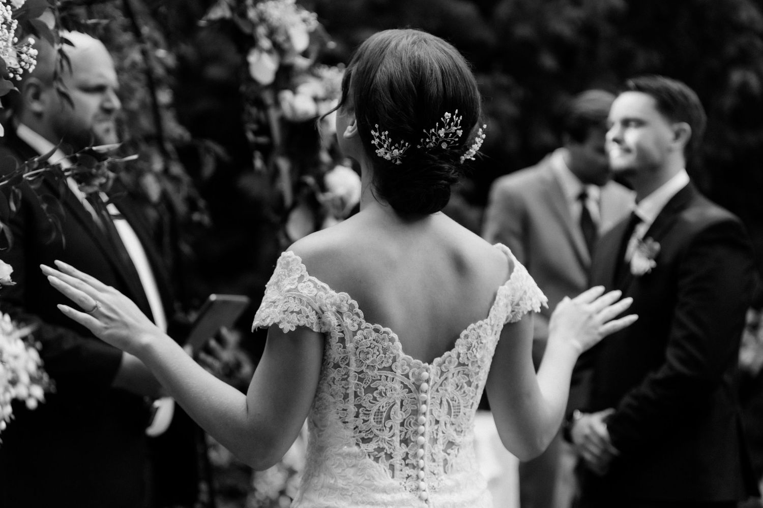 A bride in a detailed lace dress gestures during a black and white wedding ceremony moment.