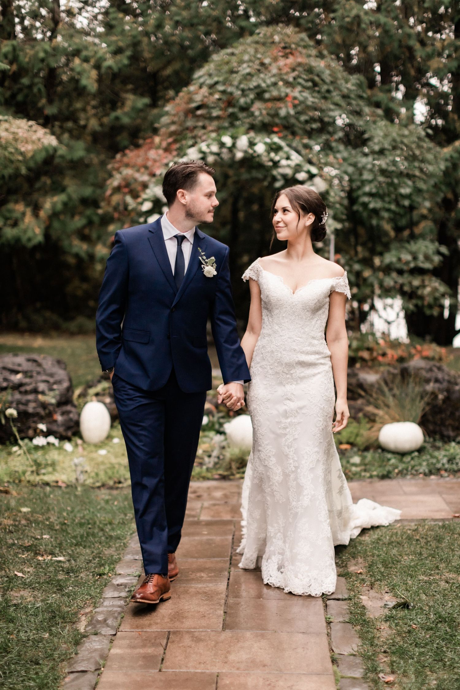 Wedding couple walking hand in hand on brick pathway with floral arch and white pumpkin decor.