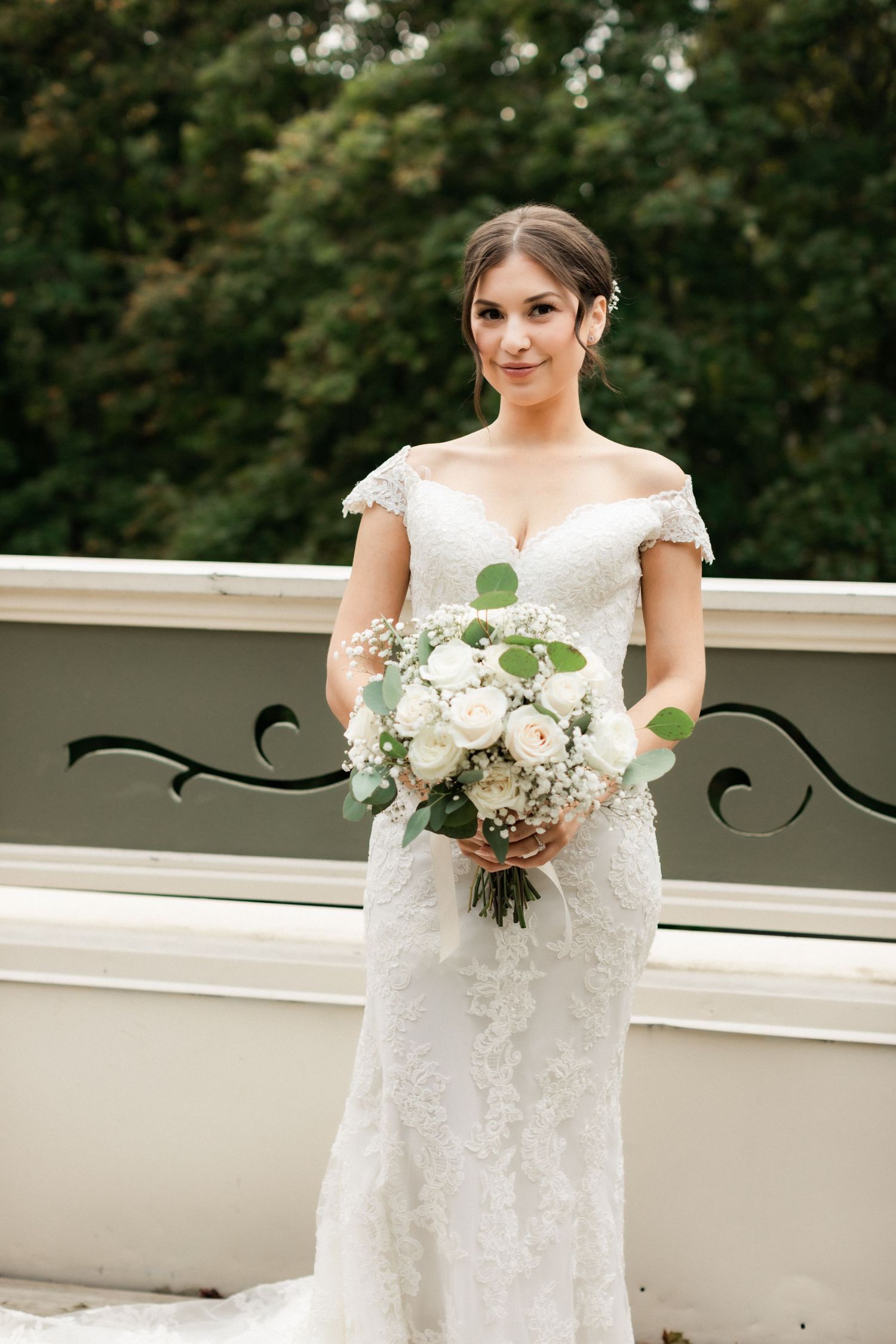 Bride poses with white rose bouquet in fitted lace wedding gown with cap sleeves on outdoor terrace.