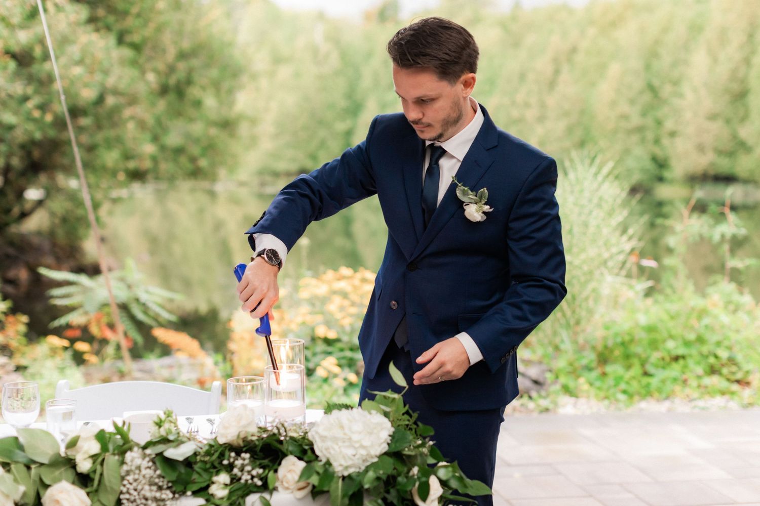 Groom in navy suit cuts wedding cake decorated with white flowers and greenery.