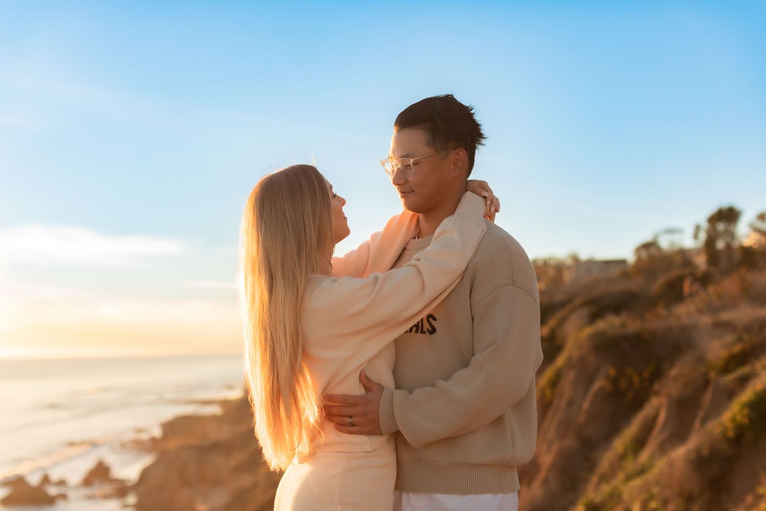 Romantic couple in light outfits sharing tender moment on coastal cliffs.