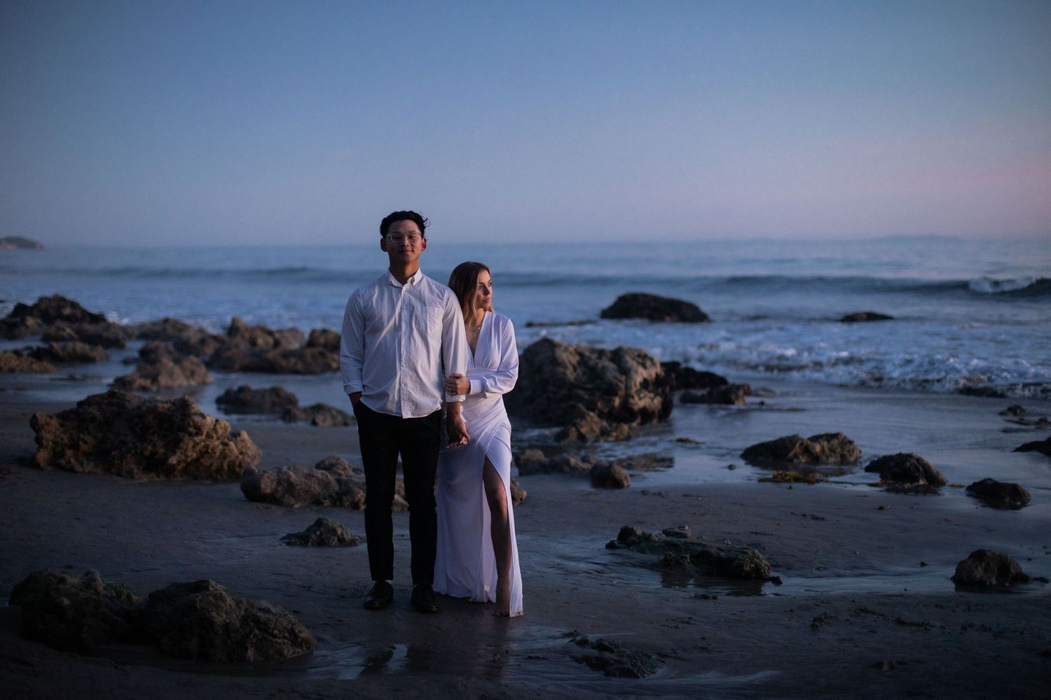 Elegant couple poses on rocky beach during blue hour photography session.