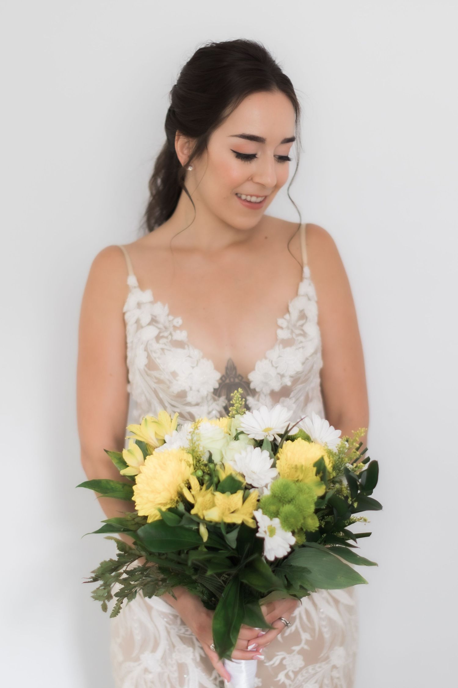 Bride in white lace gown holding fresh bouquet of yellow and white flowers with greenery.