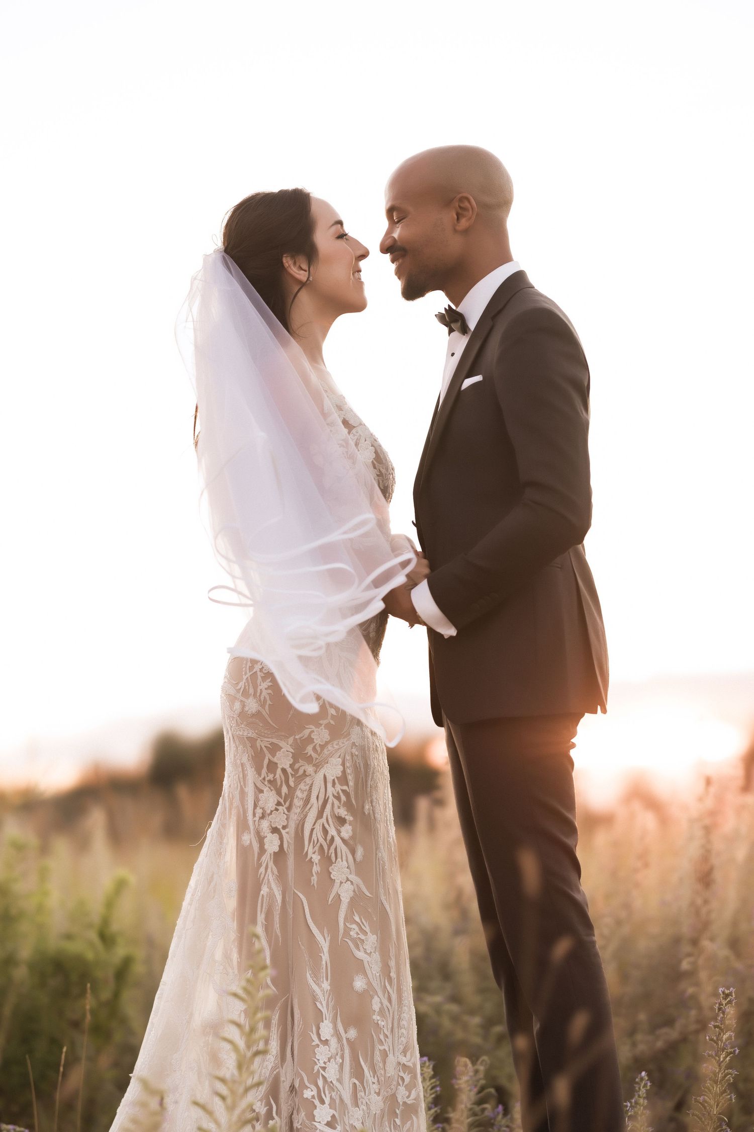 A romantic wedding couple shares an intimate moment in a field during golden hour sunset.