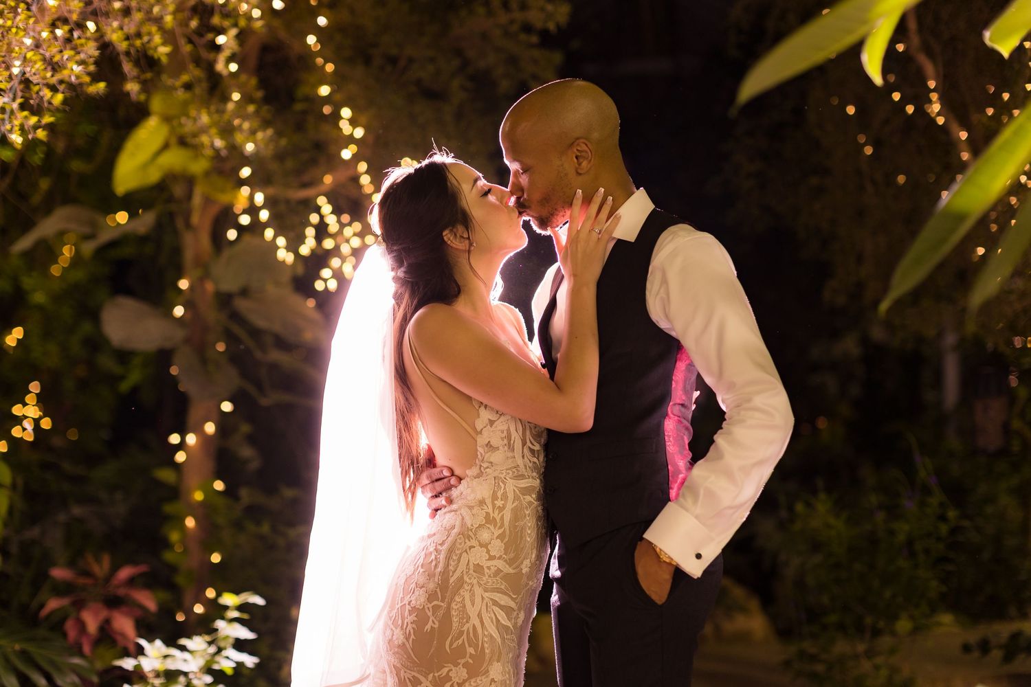Romantic nighttime portrait of newlyweds surrounded by glowing fairy lights in garden setting.