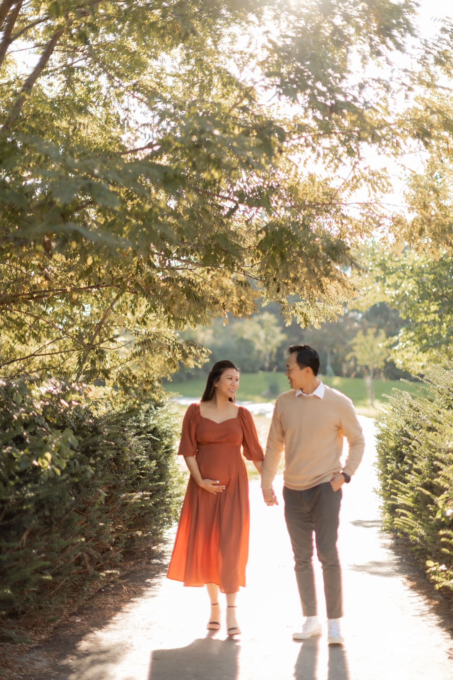 Pregnant couple walking hand in hand on sunlit path through garden during maternity shoot.