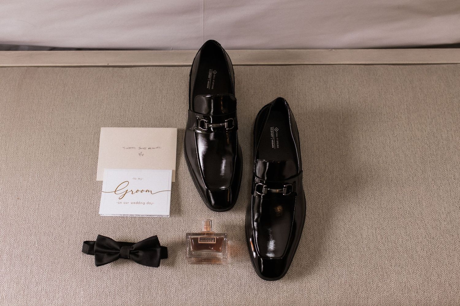 Wedding details featuring groom's black dress shoes, bow tie and letter on beige surface.