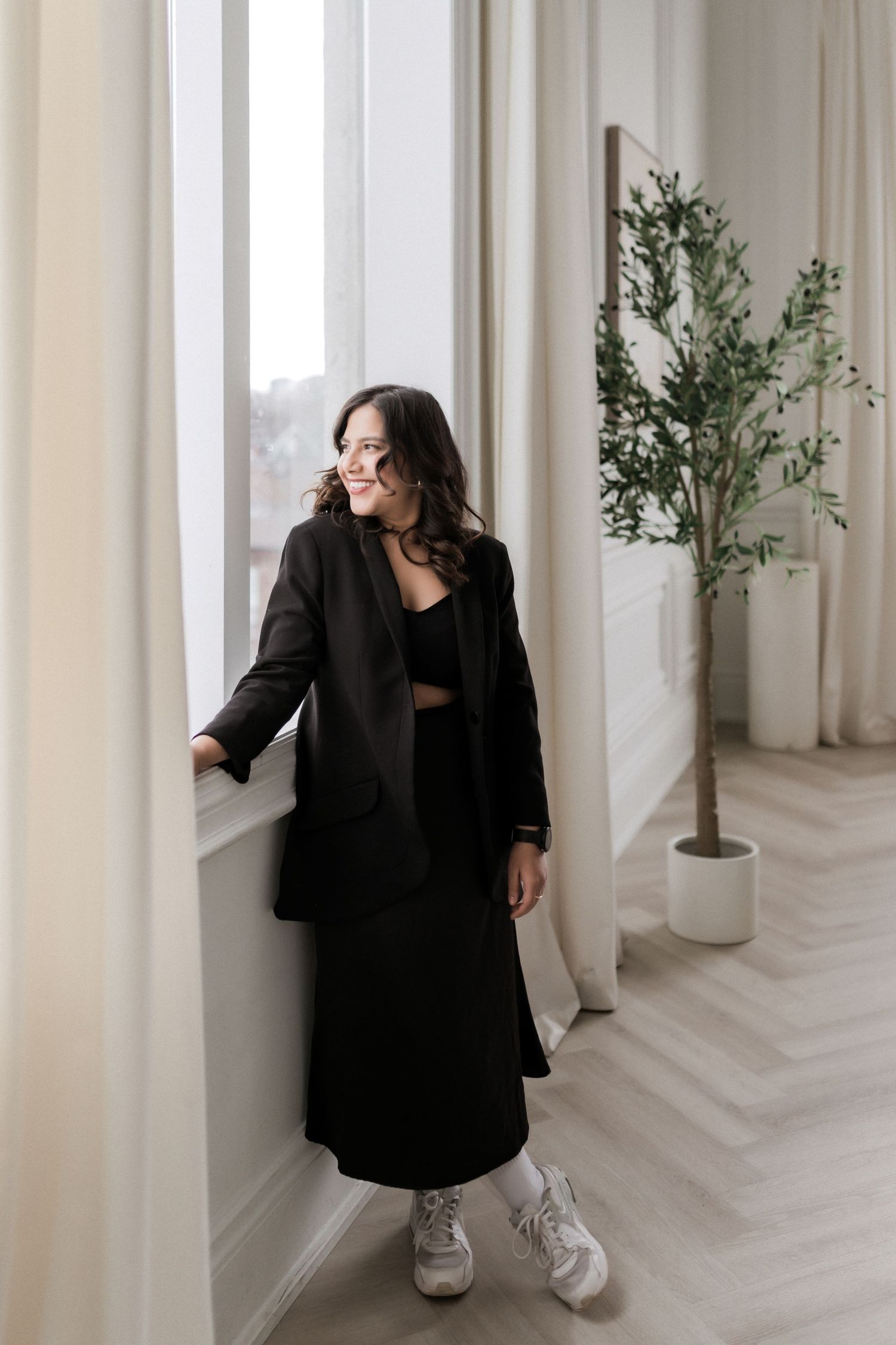 Woman in black blazer and skirt outfit looking out window in bright minimalist room.