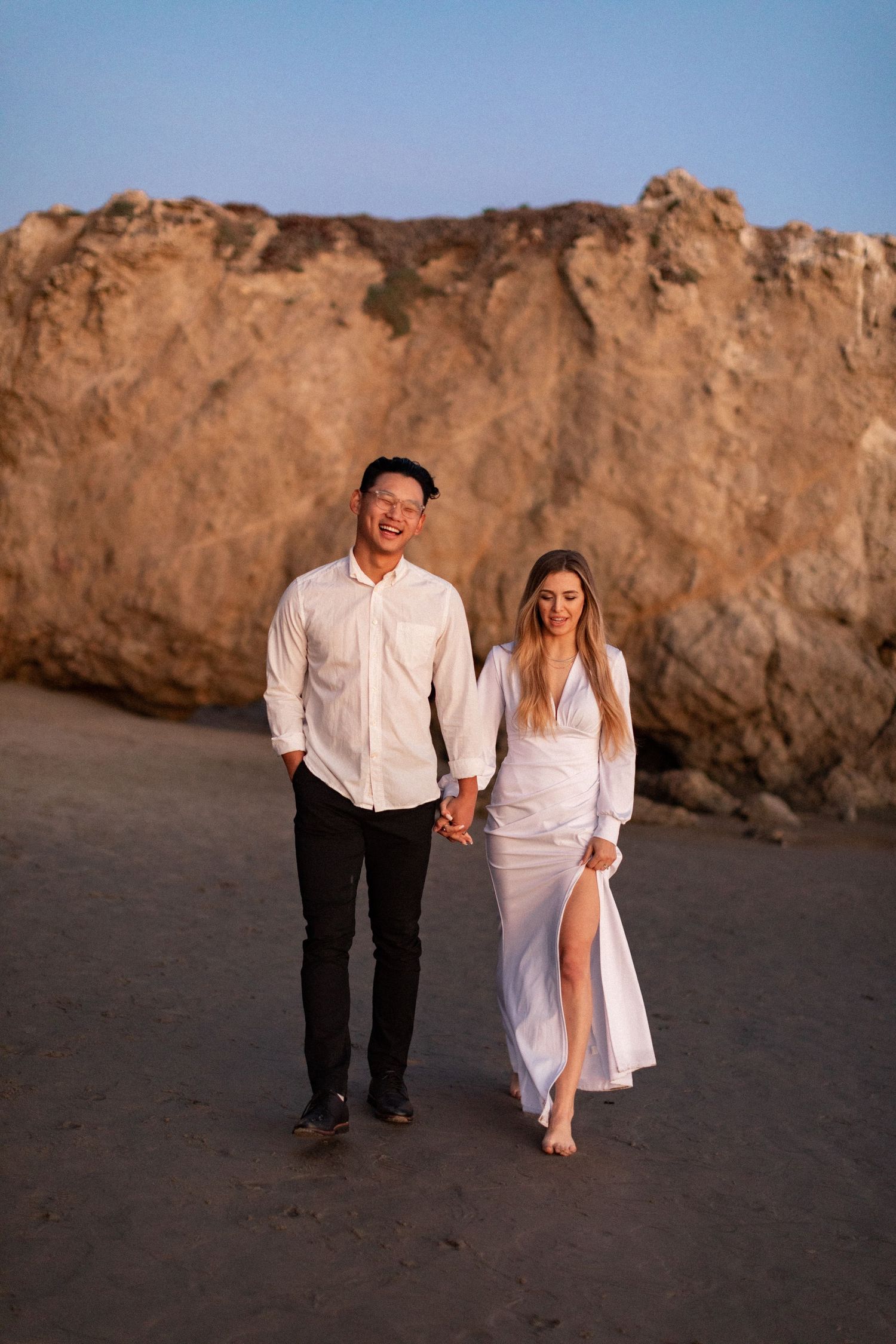 Couple in white outfits posing in front of large coastal rock formation during sunset.