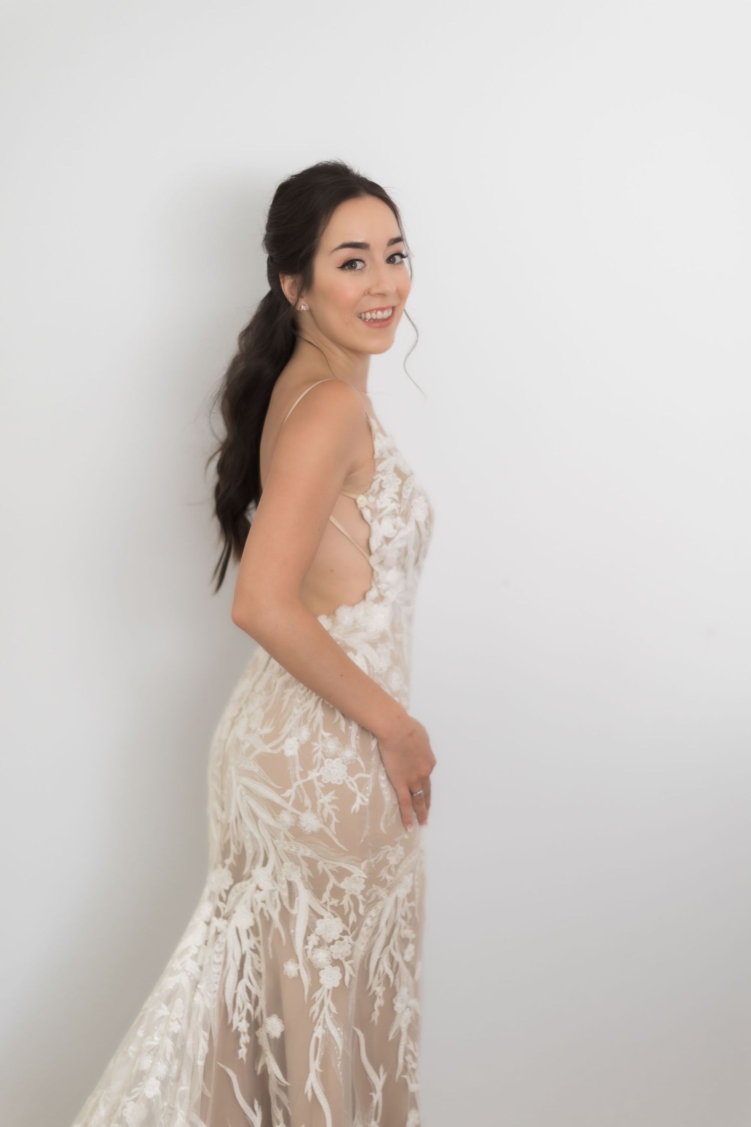Woman in backless lace wedding dress poses against white wall.