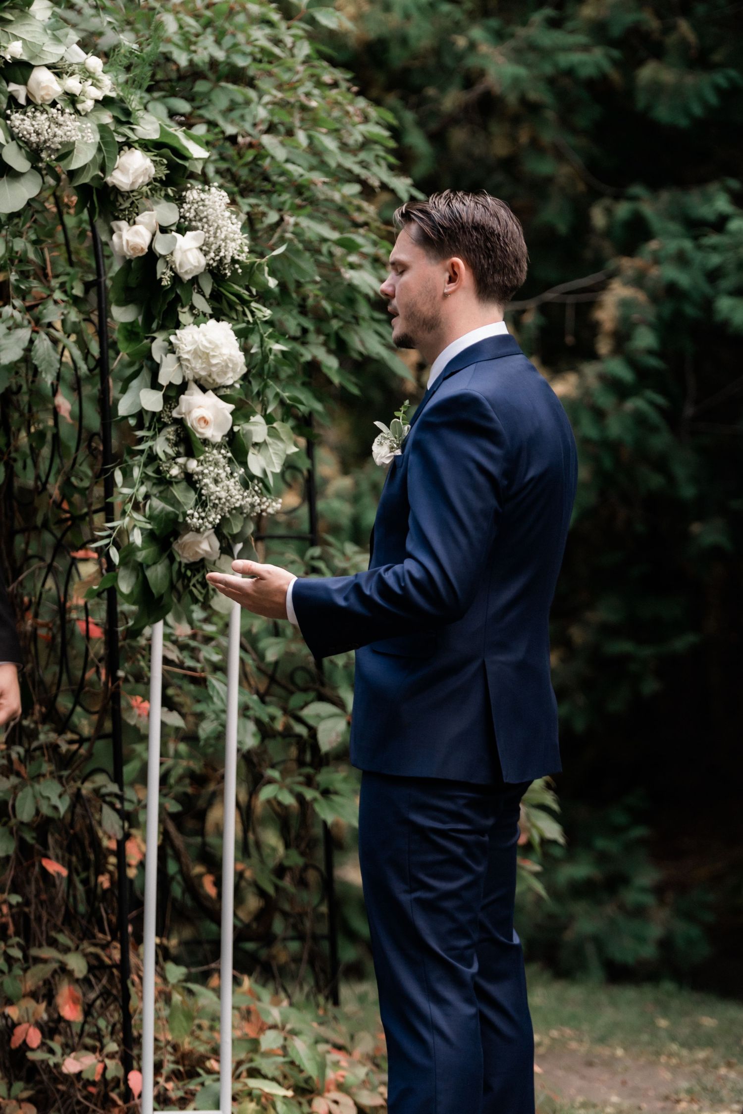 Groom in navy suit standing beside floral-decorated wedding arch in garden setting.