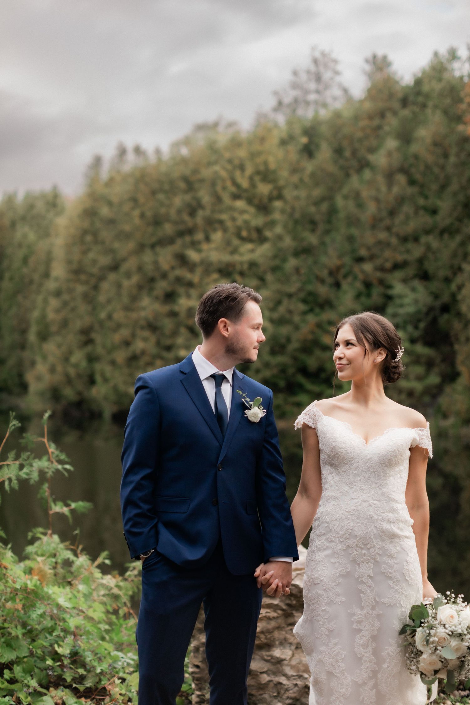 A romantic wedding portrait by the lake with elegant attire and natural forest backdrop.