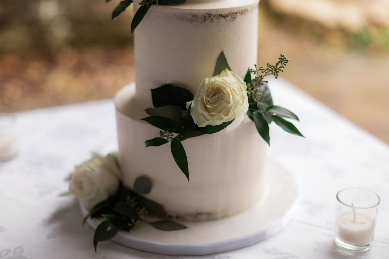 Elegant white wedding cake decorated with fresh roses and greenery on white tablecloth.